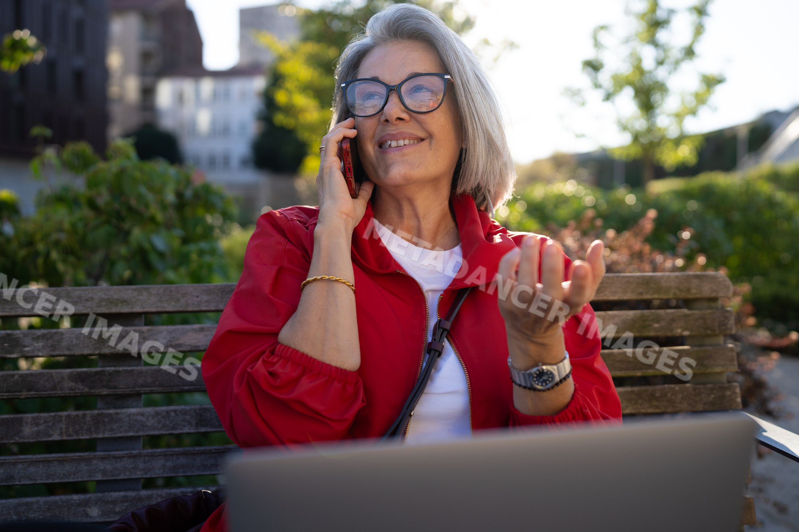 White woman talking on phone outdoors sitting on a bench with laptop