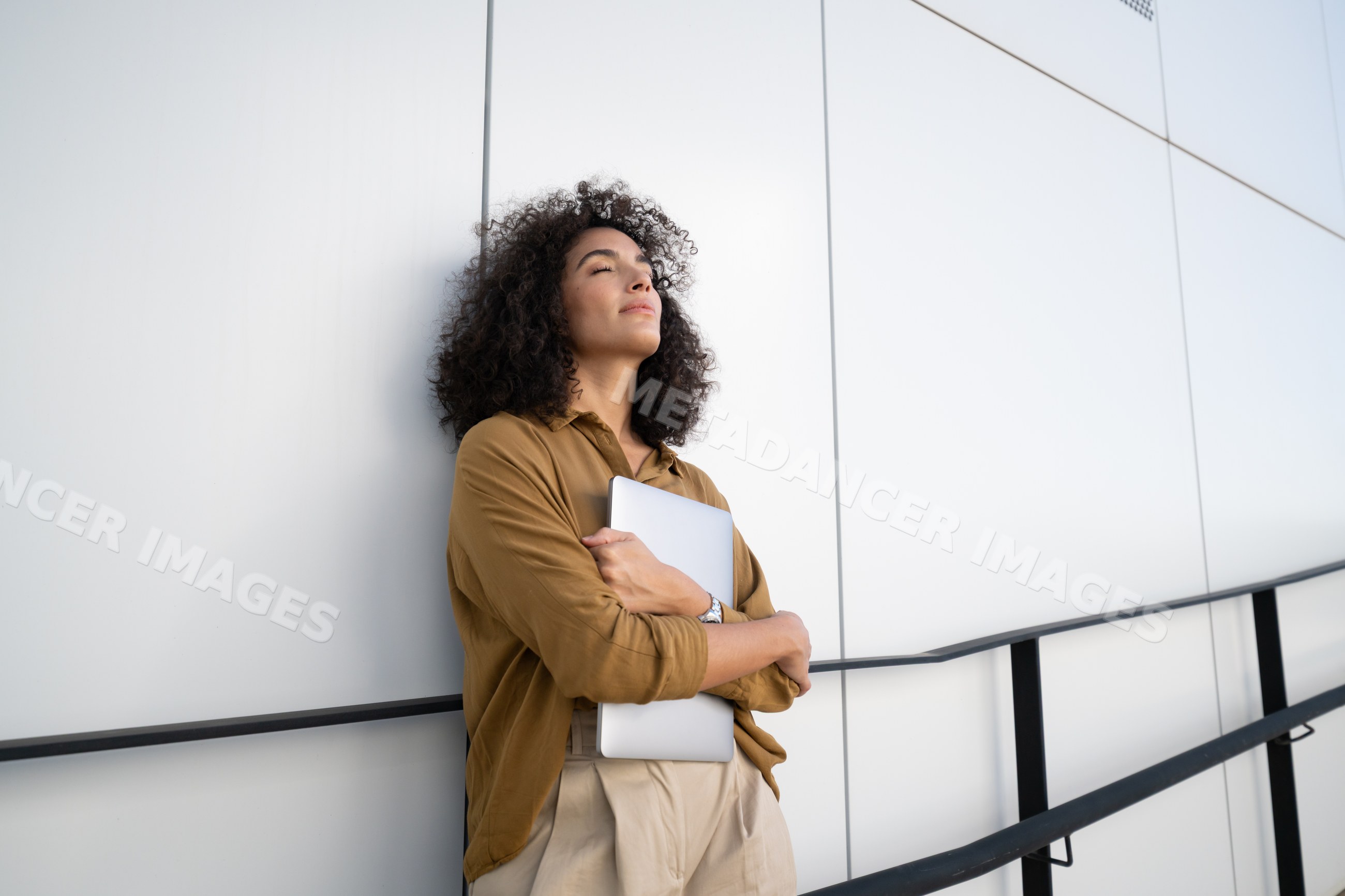 Woman holding laptop leaning against modern building wall outdoors