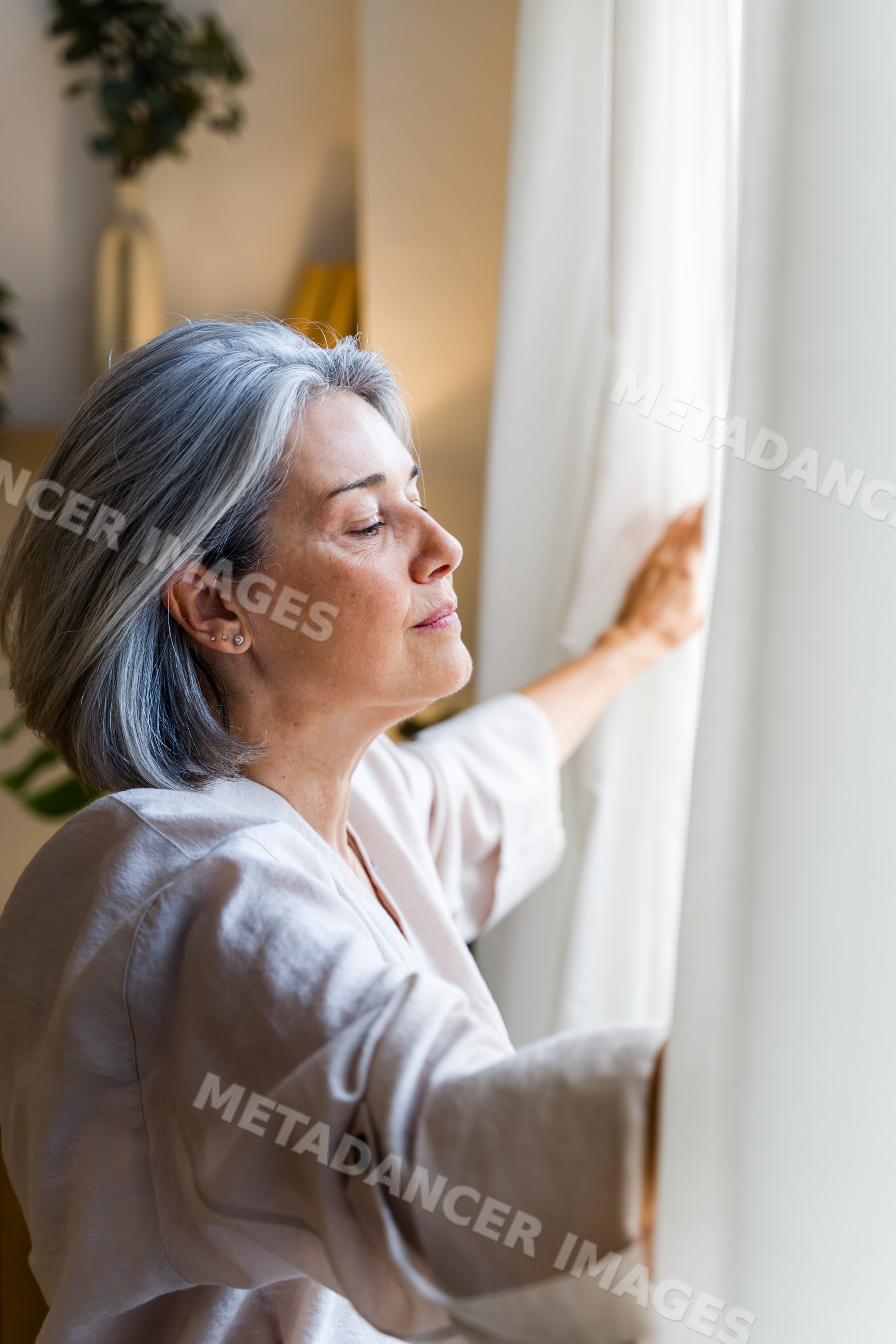 Senior white woman opening curtain to natural light indoors at home