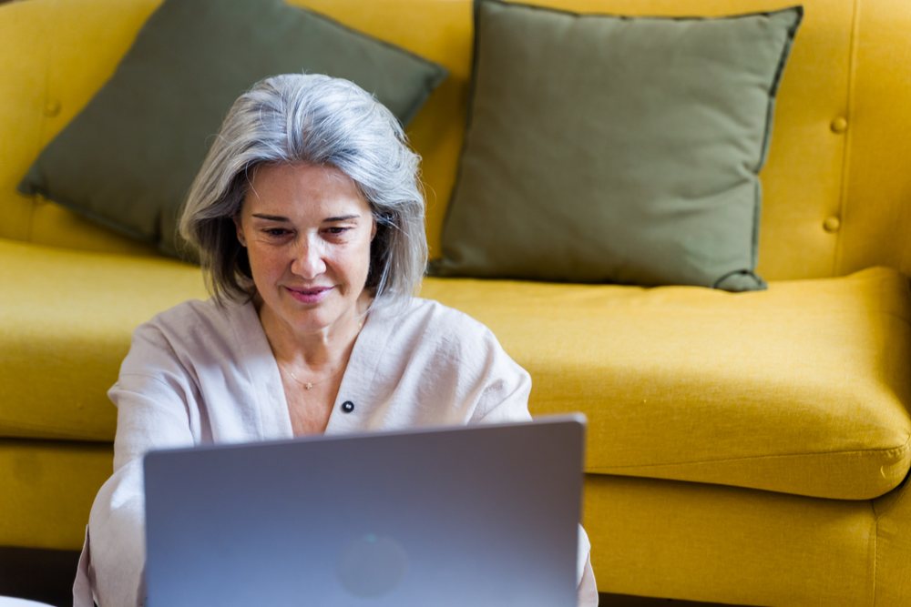 White woman sitting on floor using laptop in front of yellow sofa