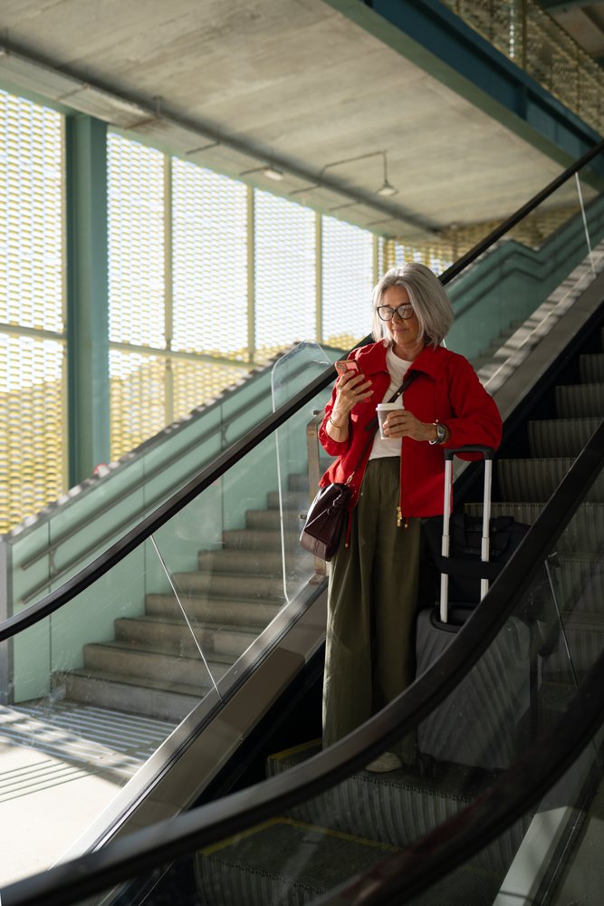 White woman with luggage stands on escalator using smartphone