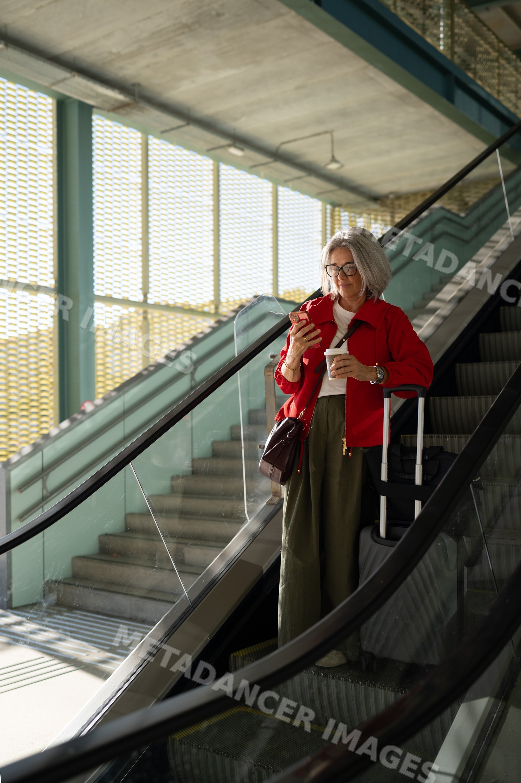 White woman with luggage stands on escalator using smartphone