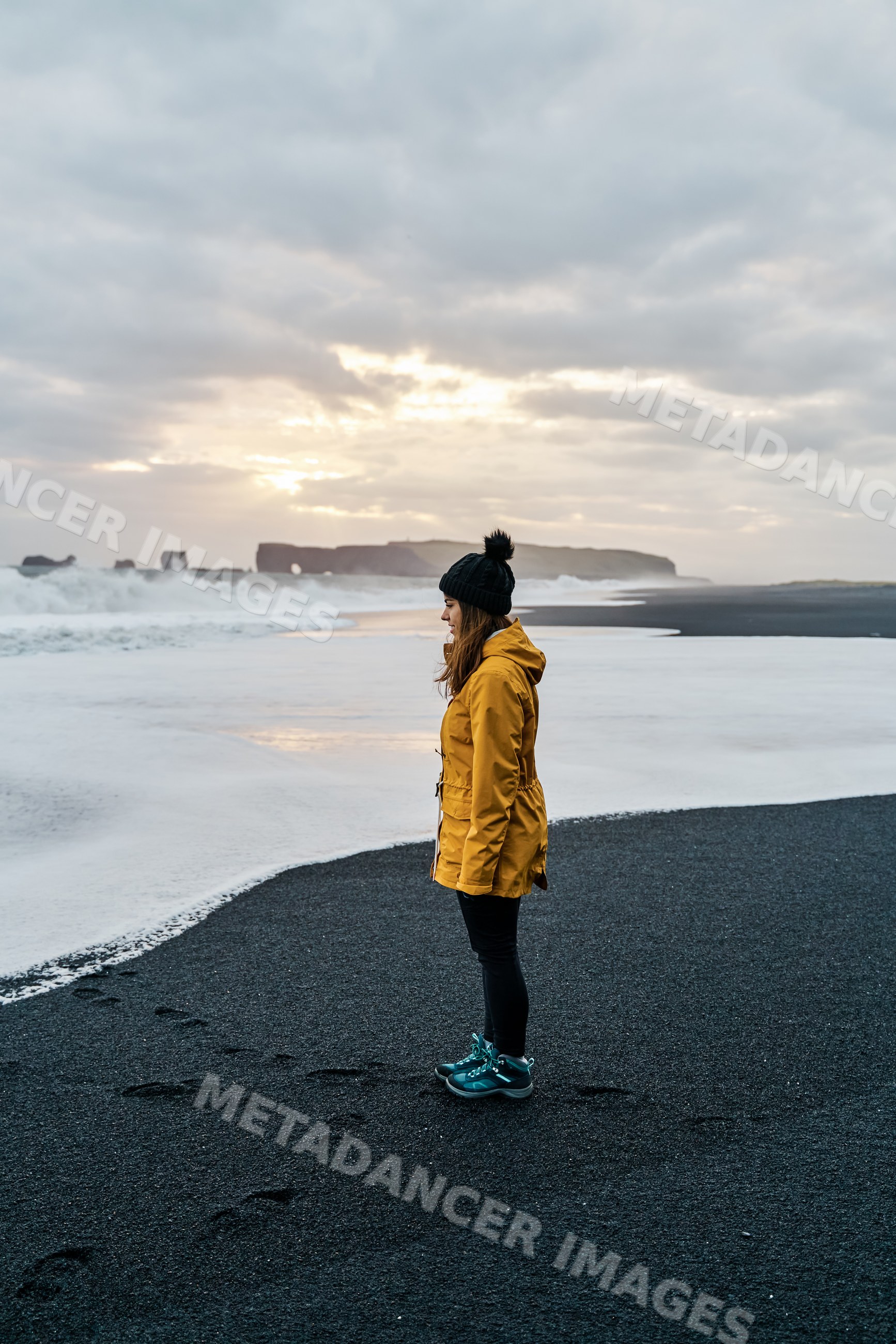 A white woman stands on a black sand beach at sunset in Iceland