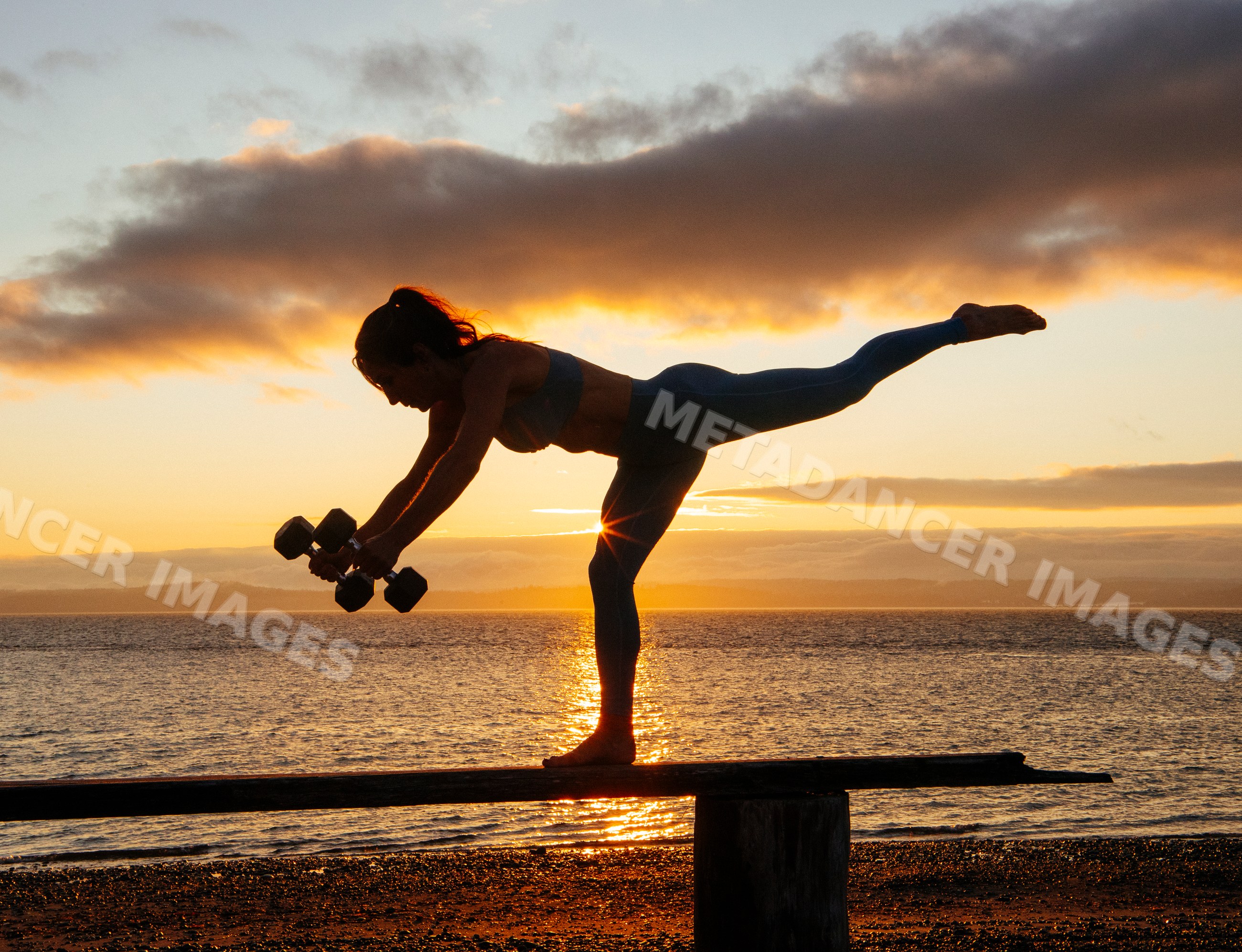 White woman balancing with dumbbells exercising on a beach at sunset