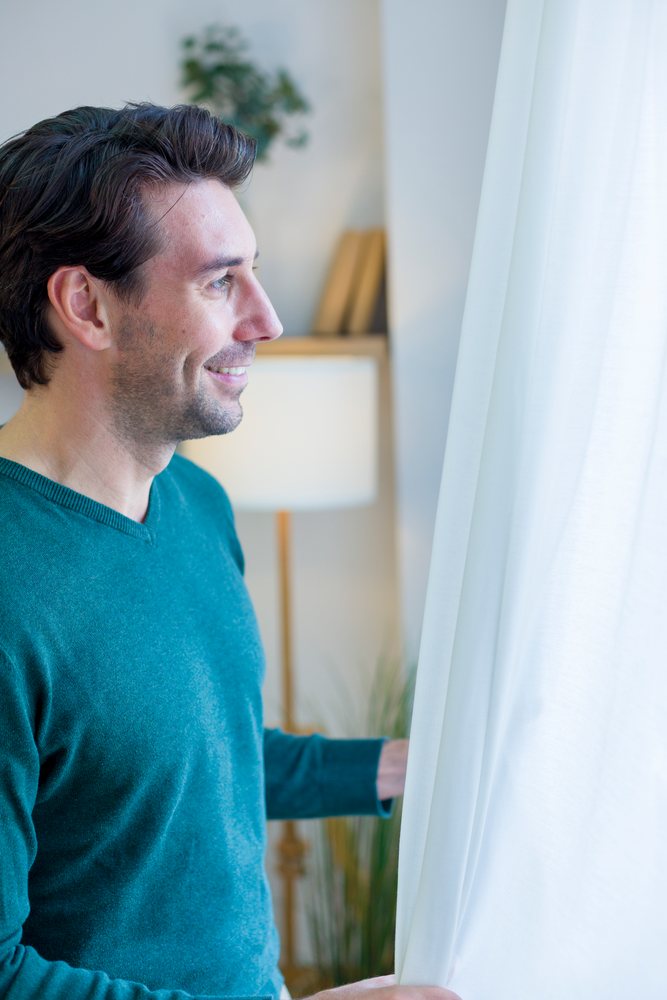 White man looking out window in cozy home interior