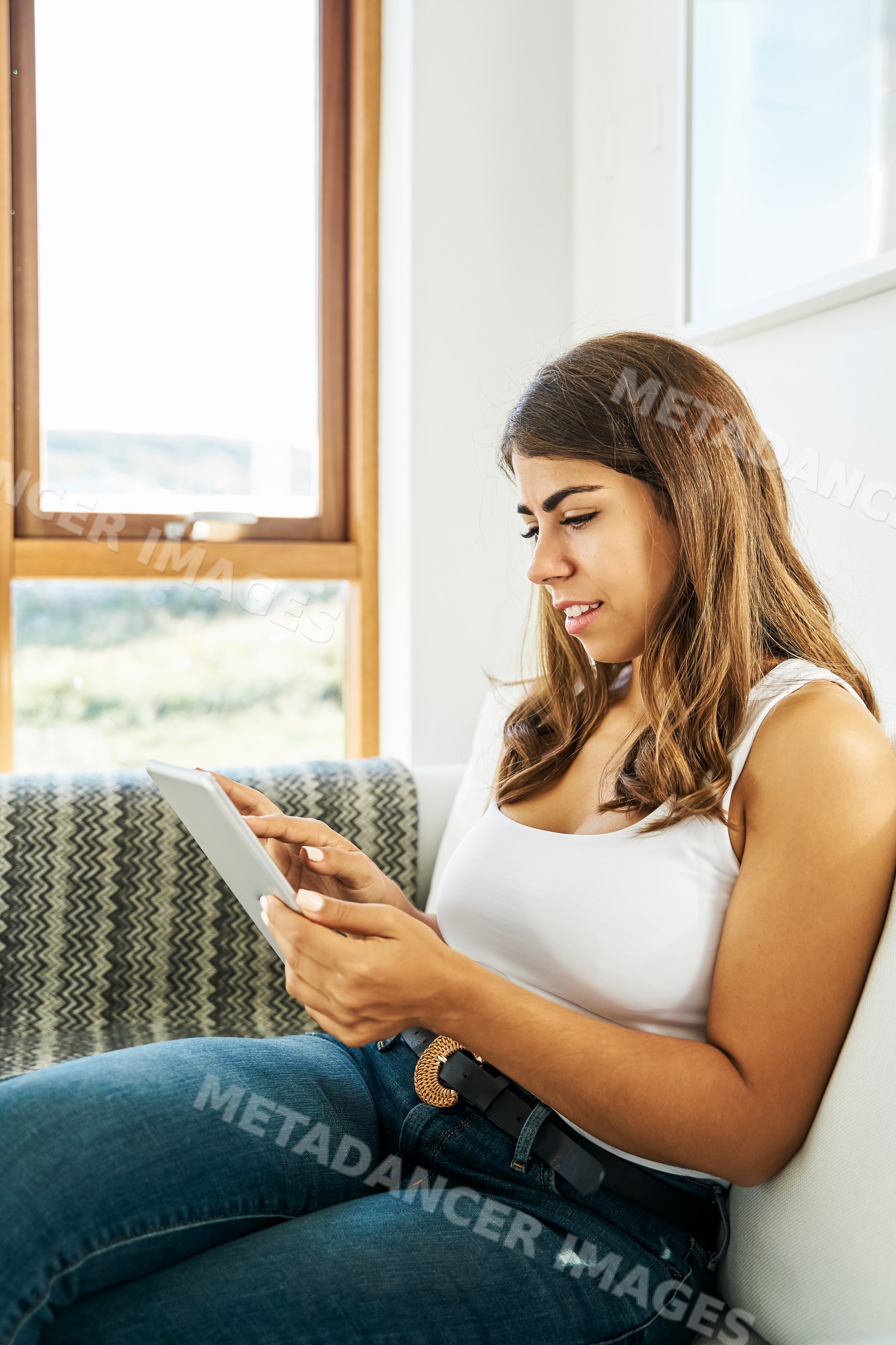 A white woman sits on a couch using a tablet in a bright home interior