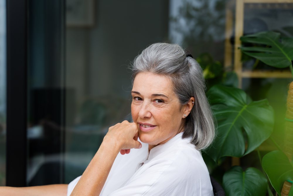 Middle-aged white woman with gray hair smiling indoors by houseplant