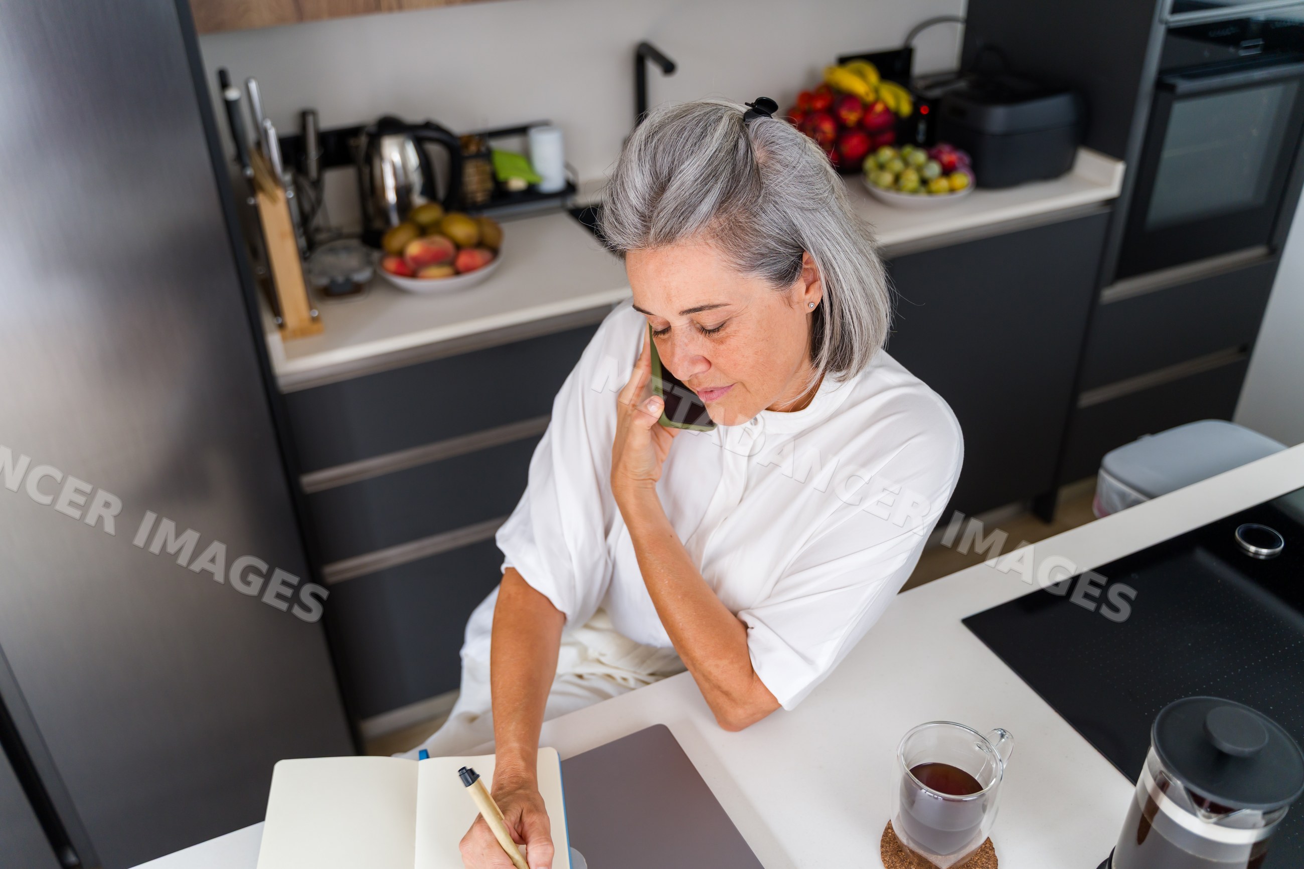 Mature white woman talking on phone and writing in kitchen in Spain