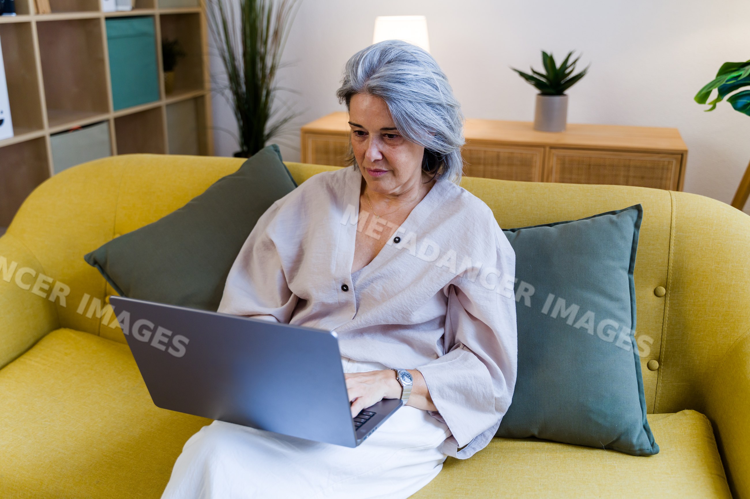 Older white woman using laptop seated on yellow couch indoors