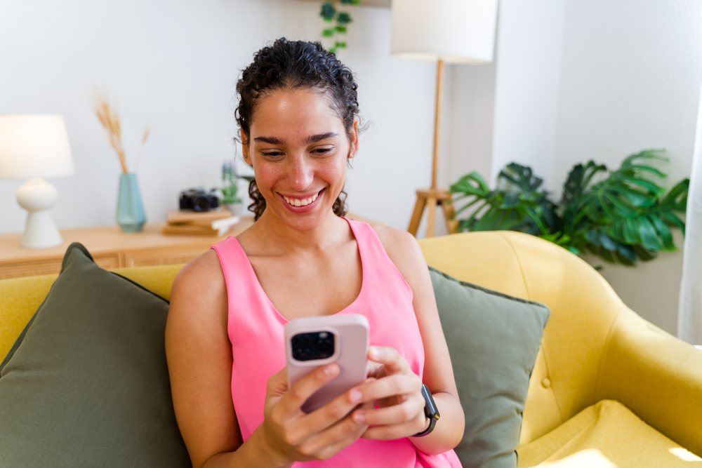 A Hispanic woman smiles while using her smartphone on a yellow sofa