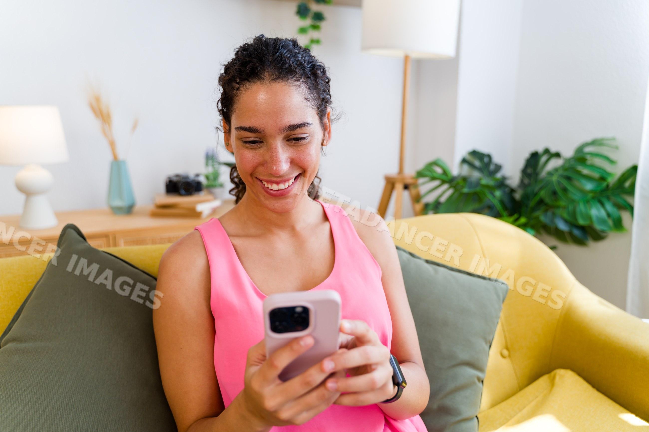 A Hispanic woman smiles while using her smartphone on a yellow sofa