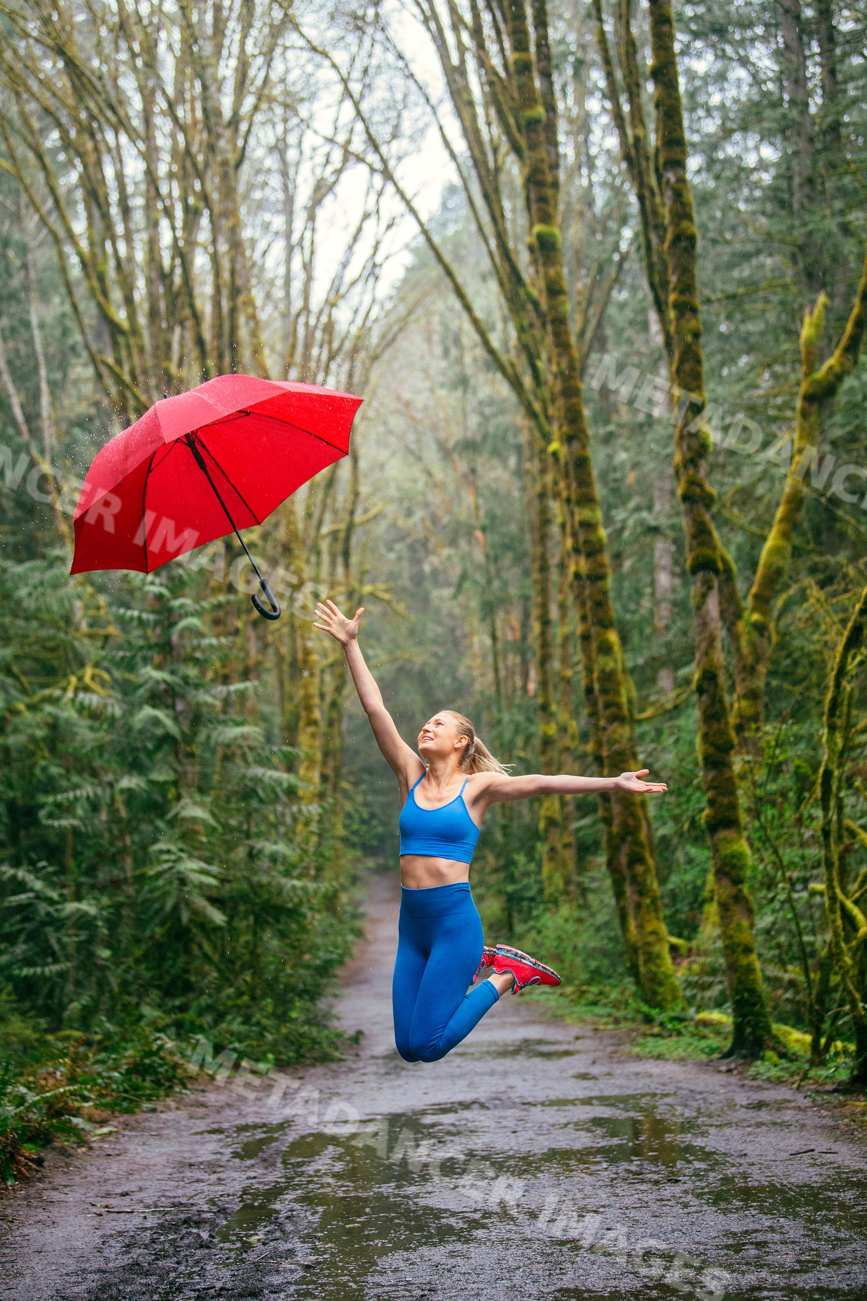 White woman jumping on rainy forest trail tossing red umbrella