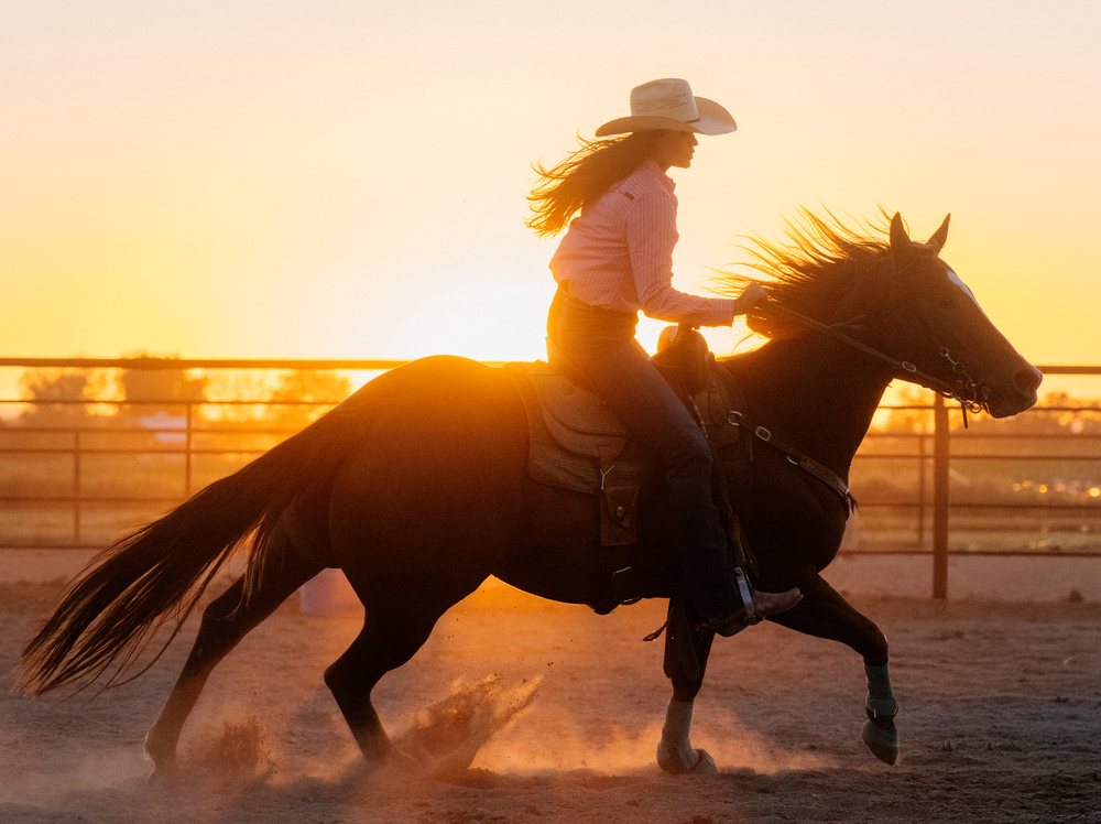 White woman horseback riding horse at sunset in outdoor arena