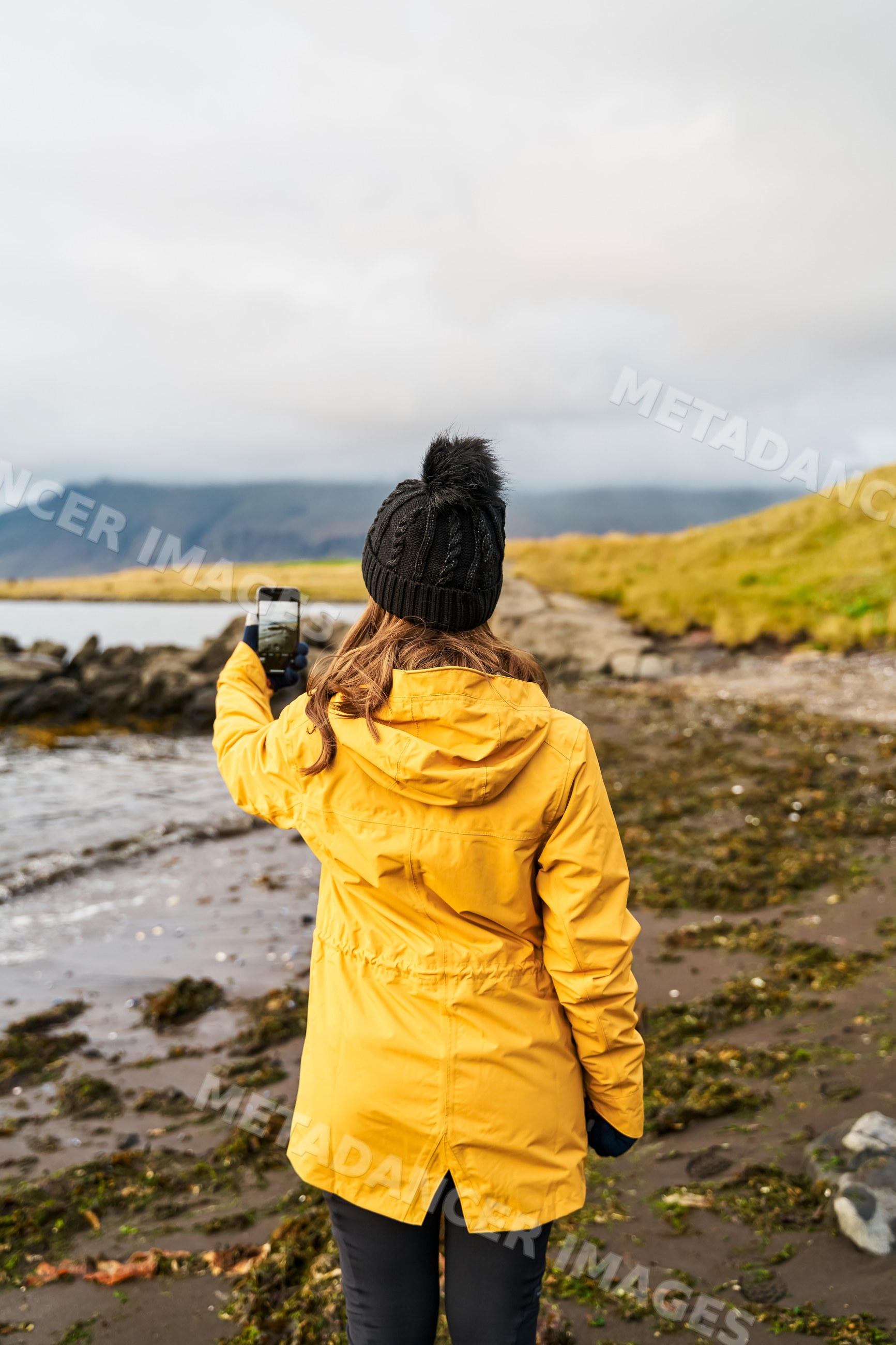 A white woman takes a smartphone photo on a rocky shoreline in a beach