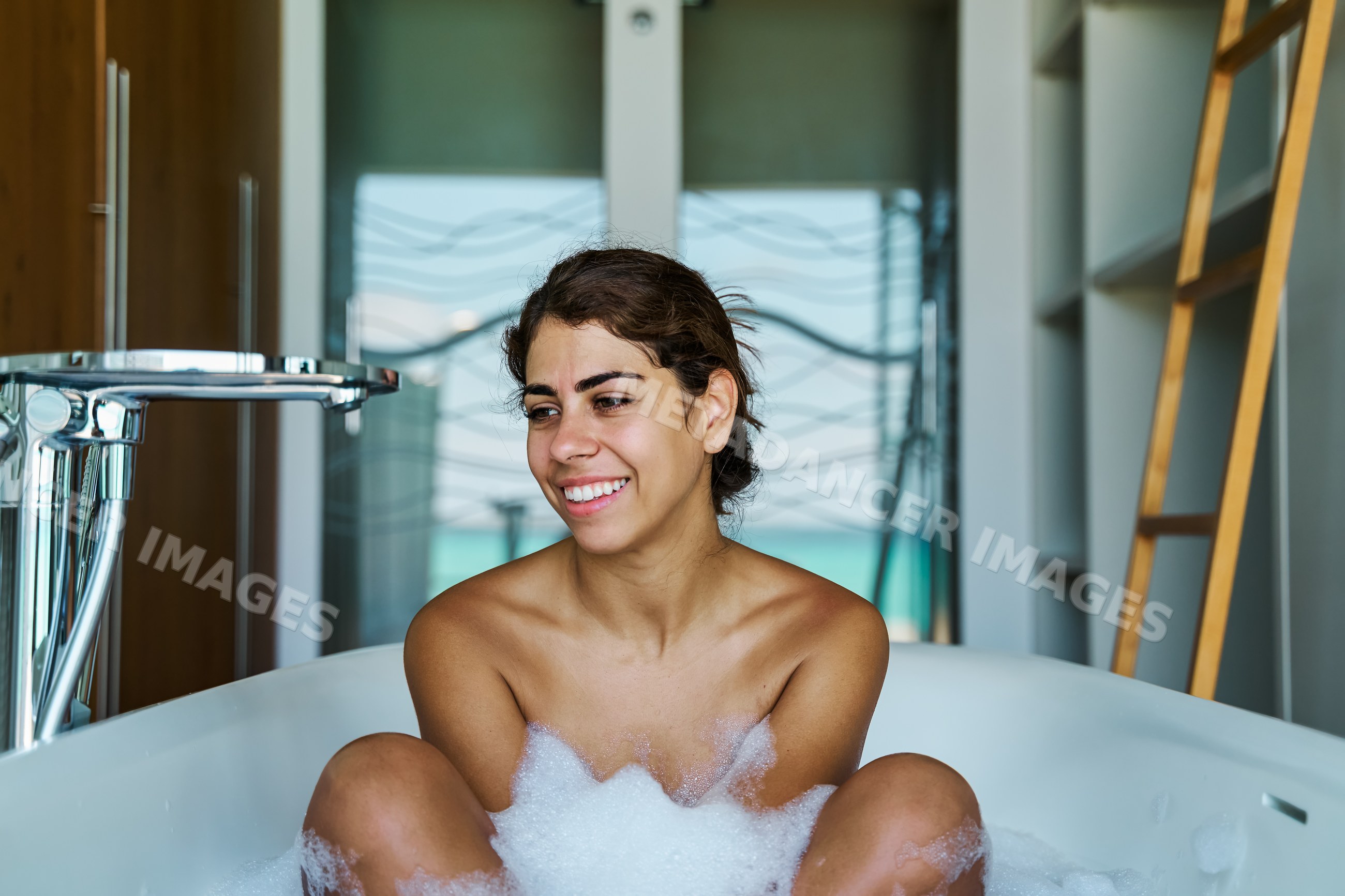 A white woman relaxing and smiling in a bubble bath inside a bathroom