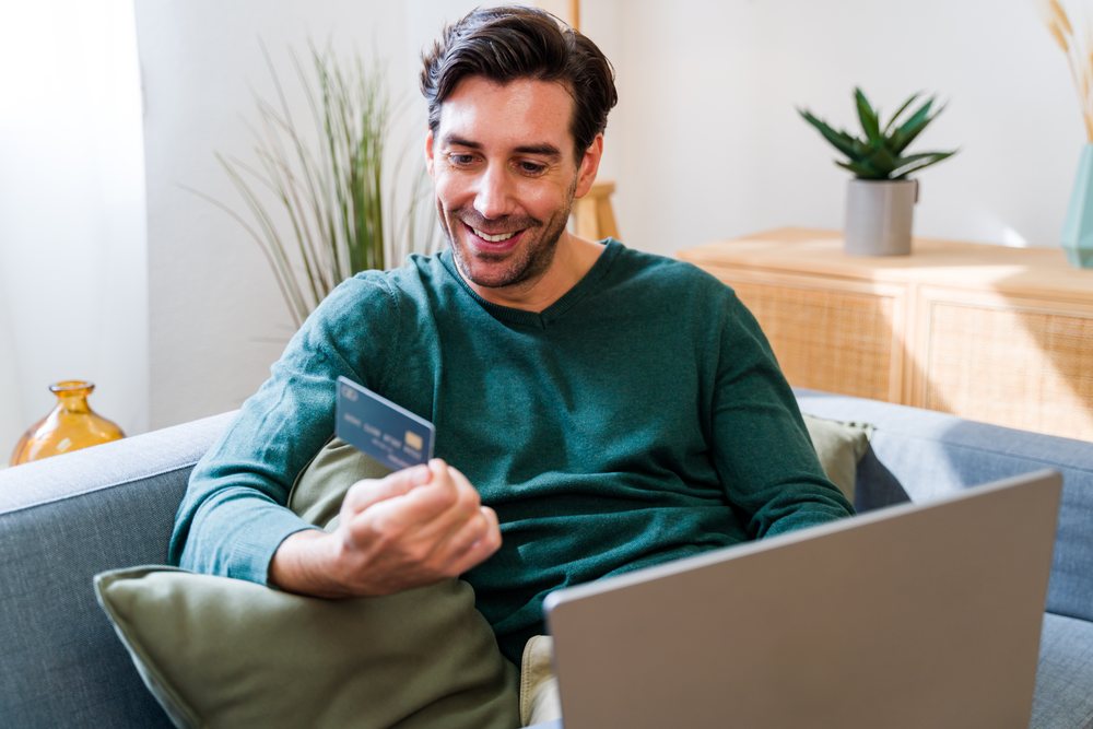 White man holding credit card using laptop in living room
