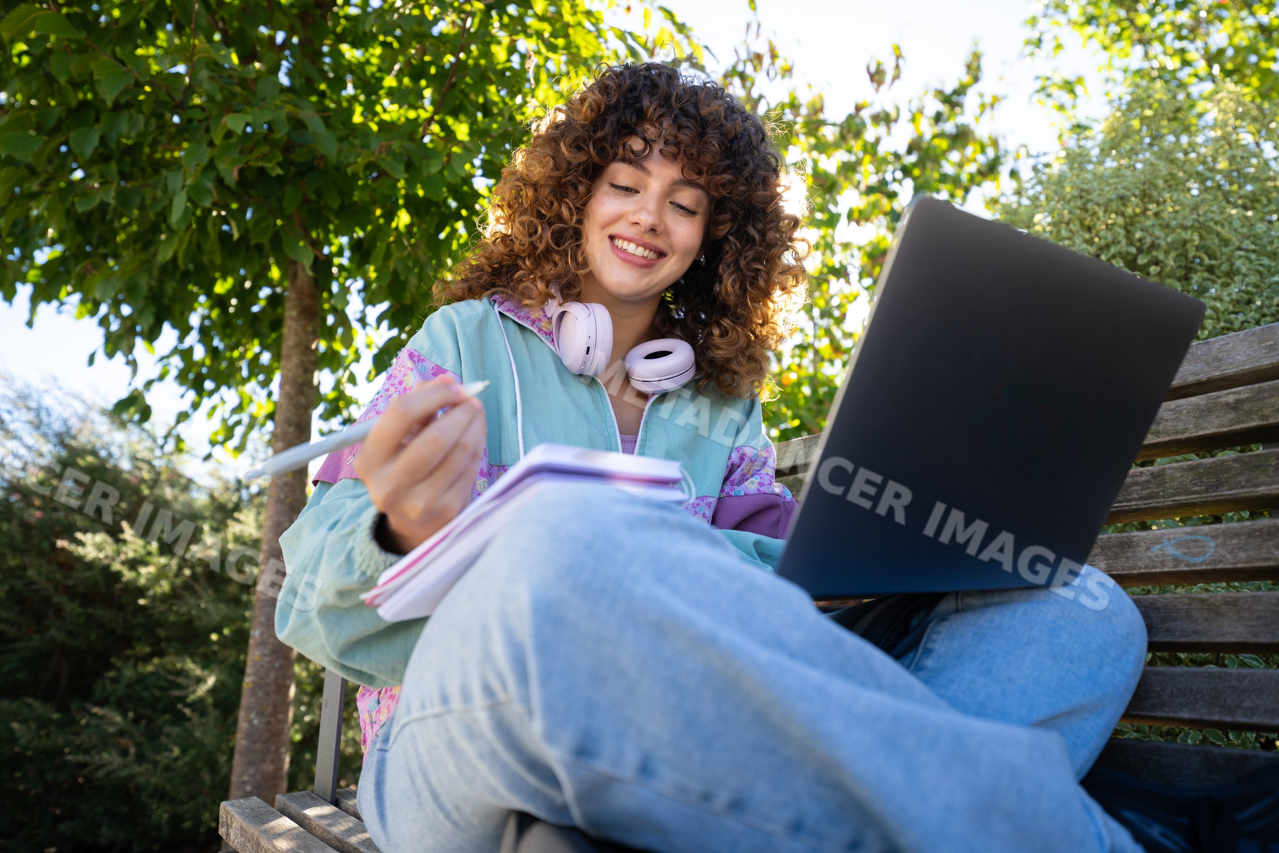 White woman writing in notebook using laptop on park bench outdoors
