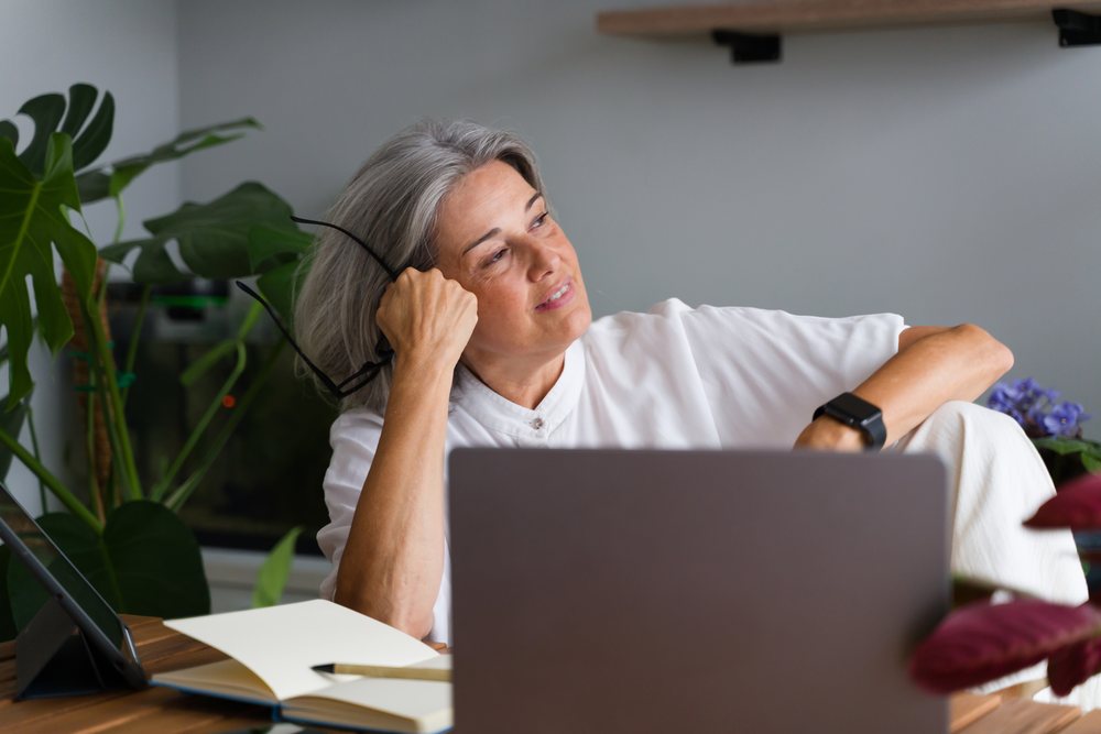 Mature white woman thoughtfully sitting with laptop and notebook