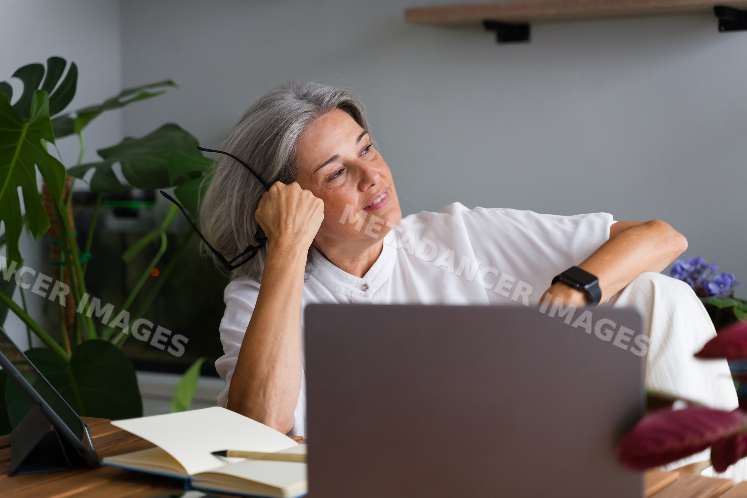 Mature white woman thoughtfully sitting with laptop and notebook