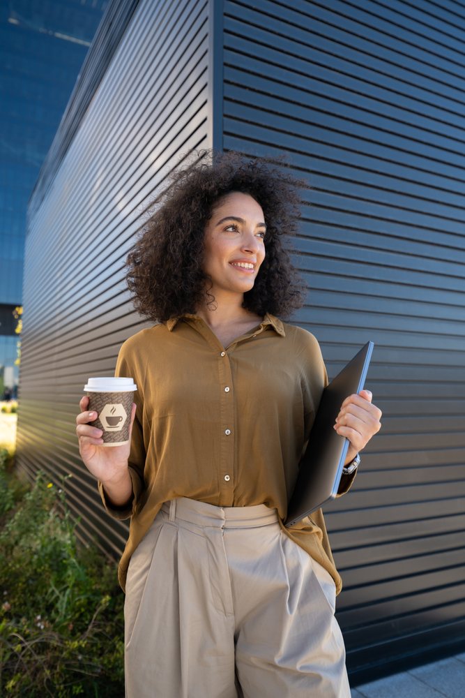 Woman holding coffee cup and laptop outdoors by modern building