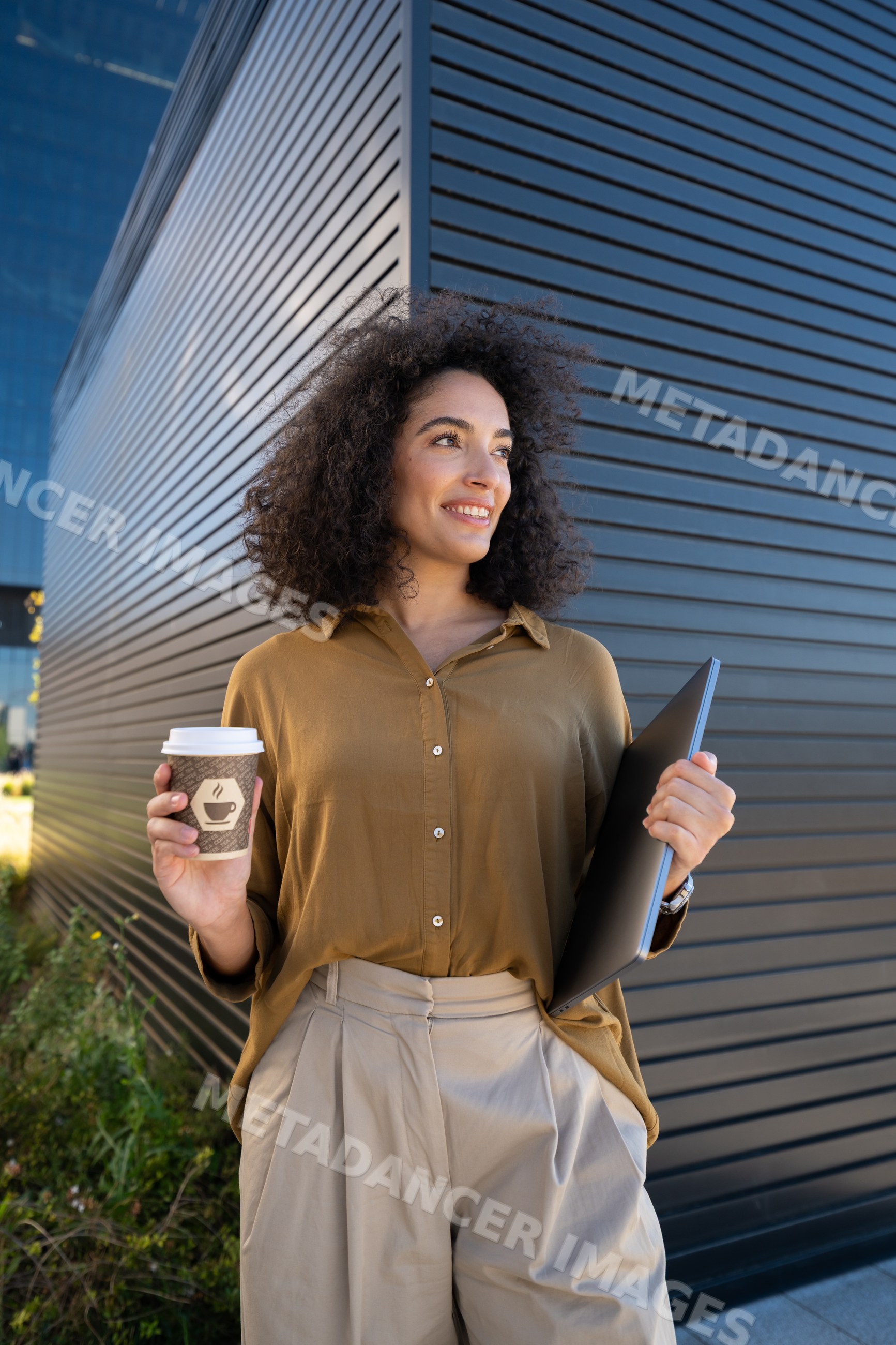 Woman holding coffee cup and laptop outdoors by modern building