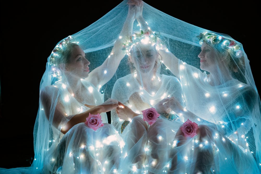 Three white women illuminated by fairy lights under veils at night