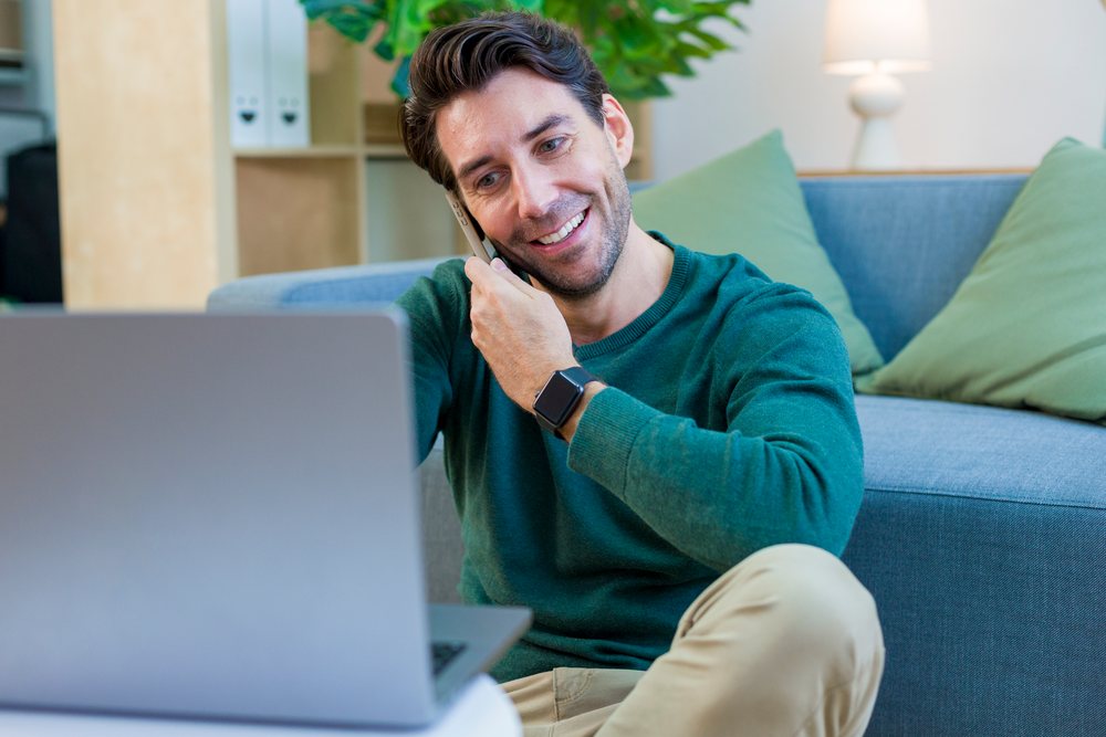 White man on smartphone call using laptop in home living room
