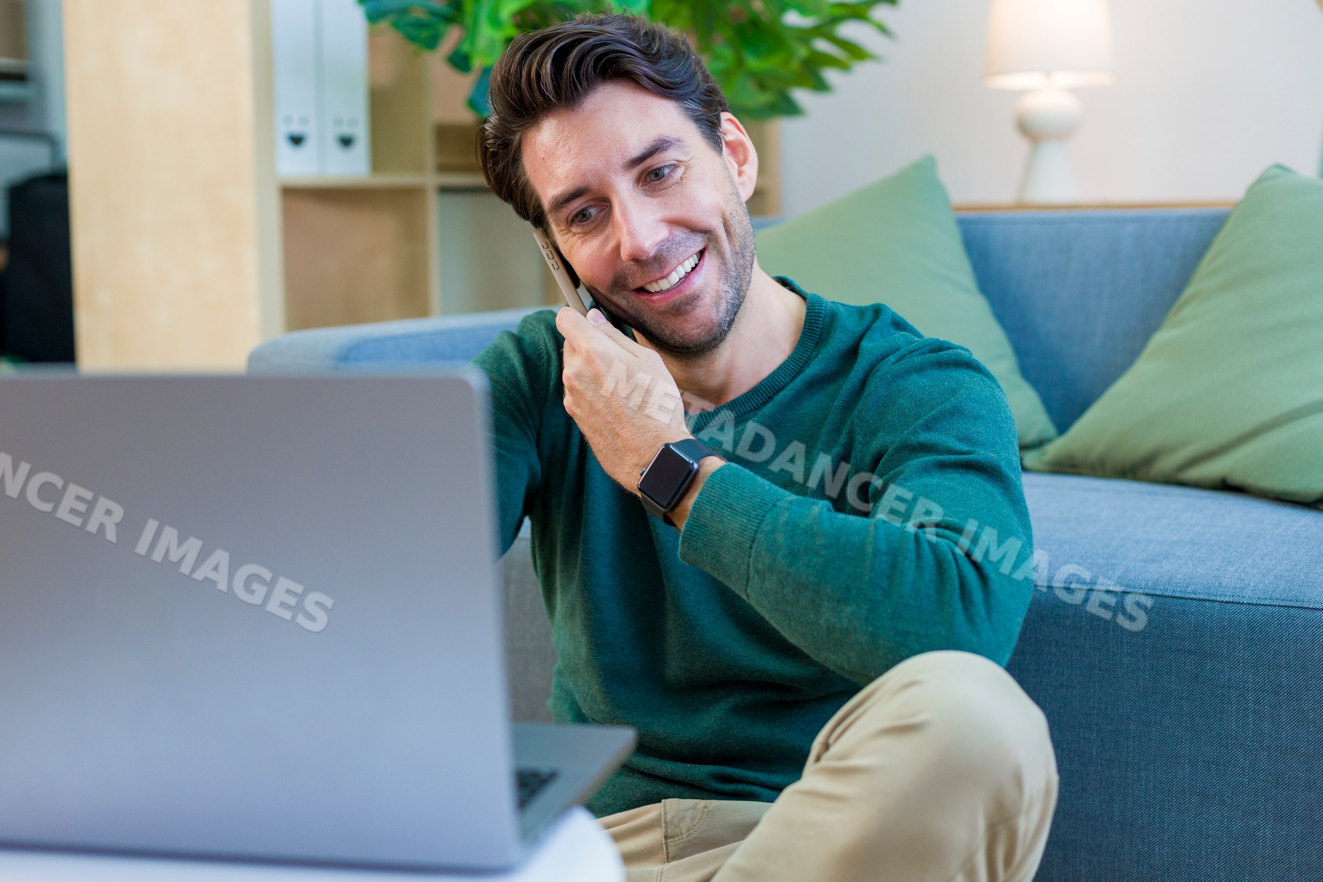 White man on smartphone call using laptop in home living room