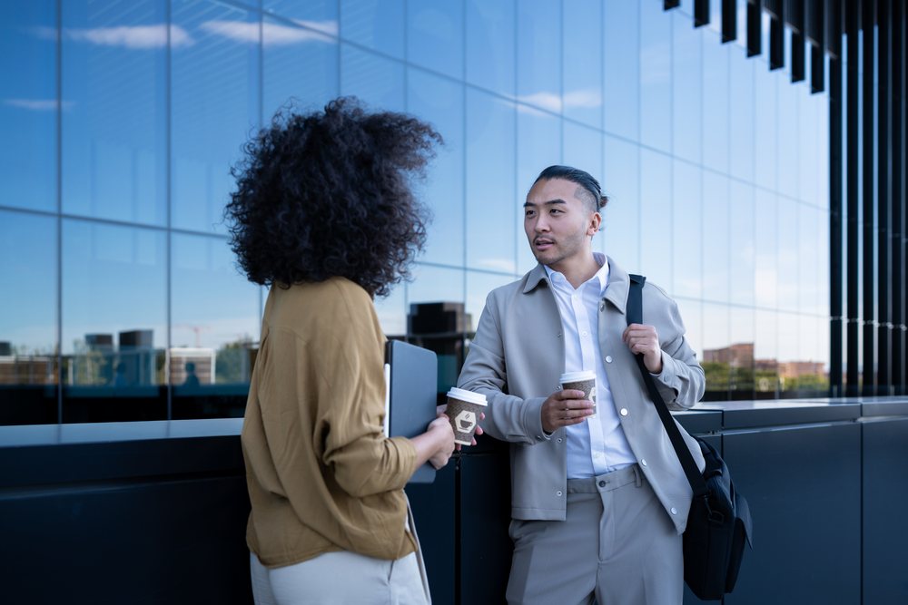 Two professionals conversing outdoors by office building with coffee