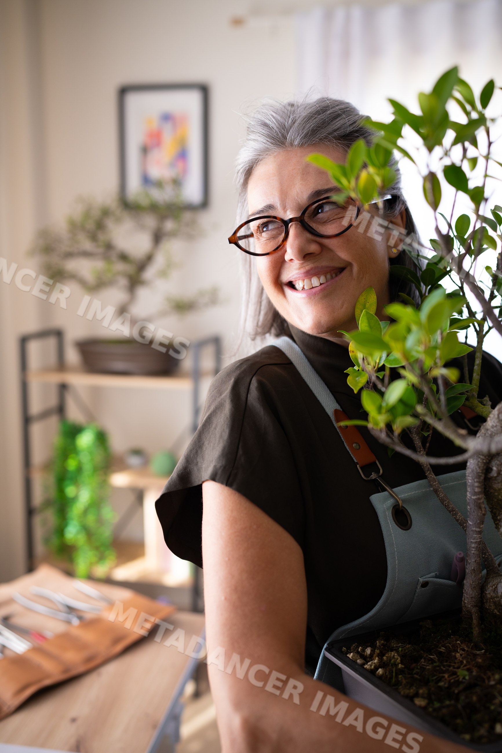 White woman holding bonsai plant indoors with gardening tools
