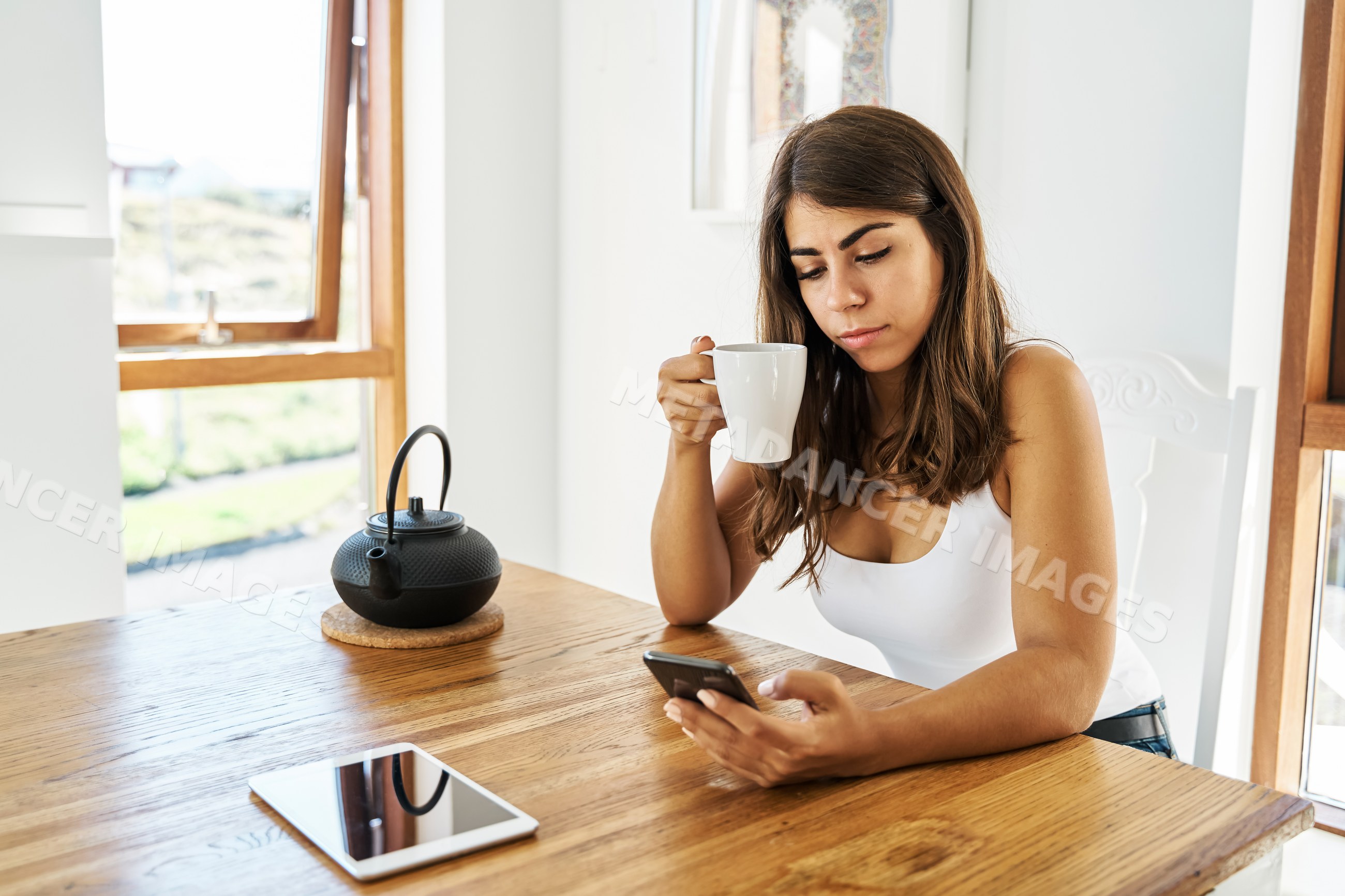 A white woman drinks tea while using her smartphone indoors