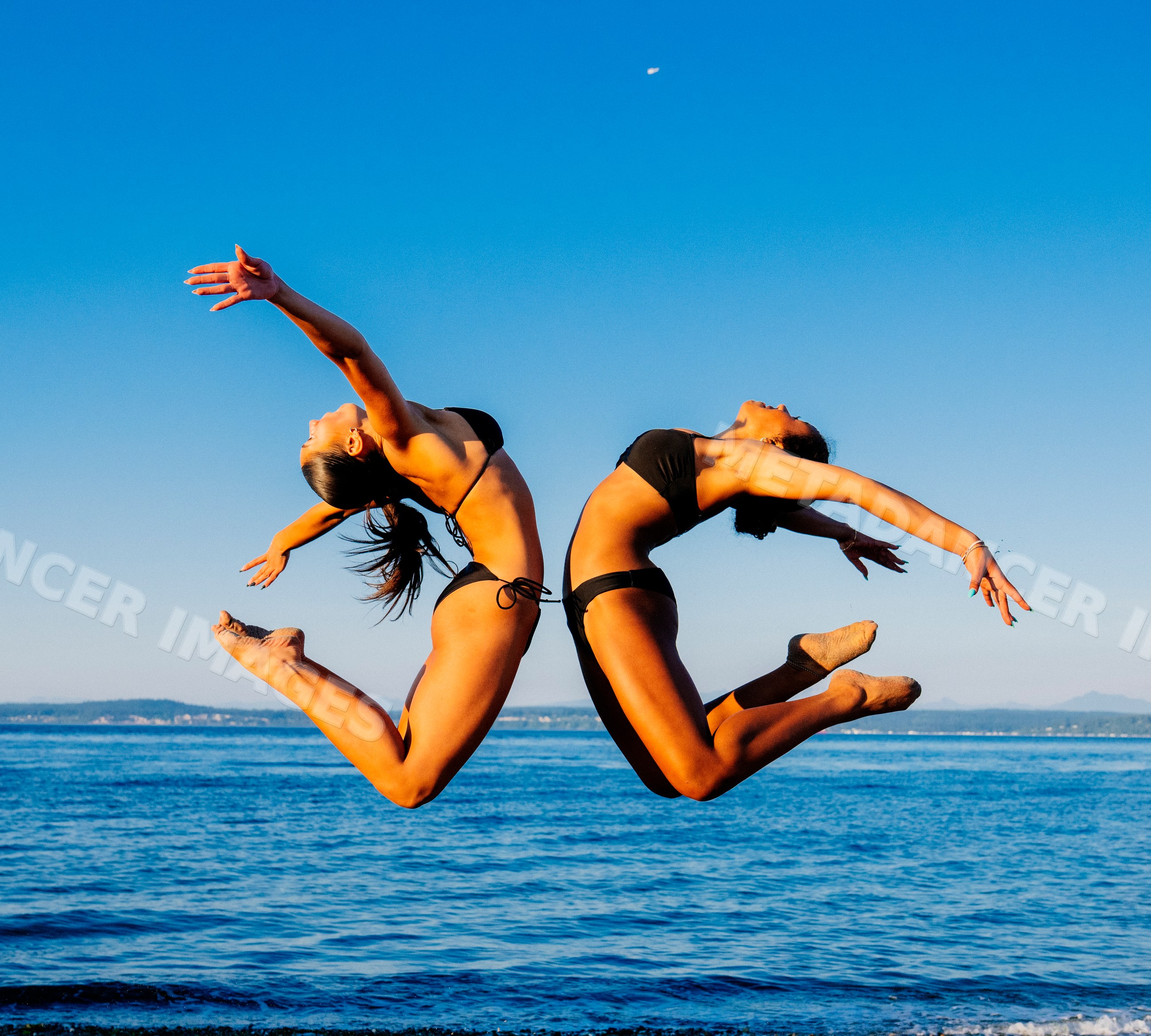 Two women in bikinis leap in synchronized backbend poses on a beach