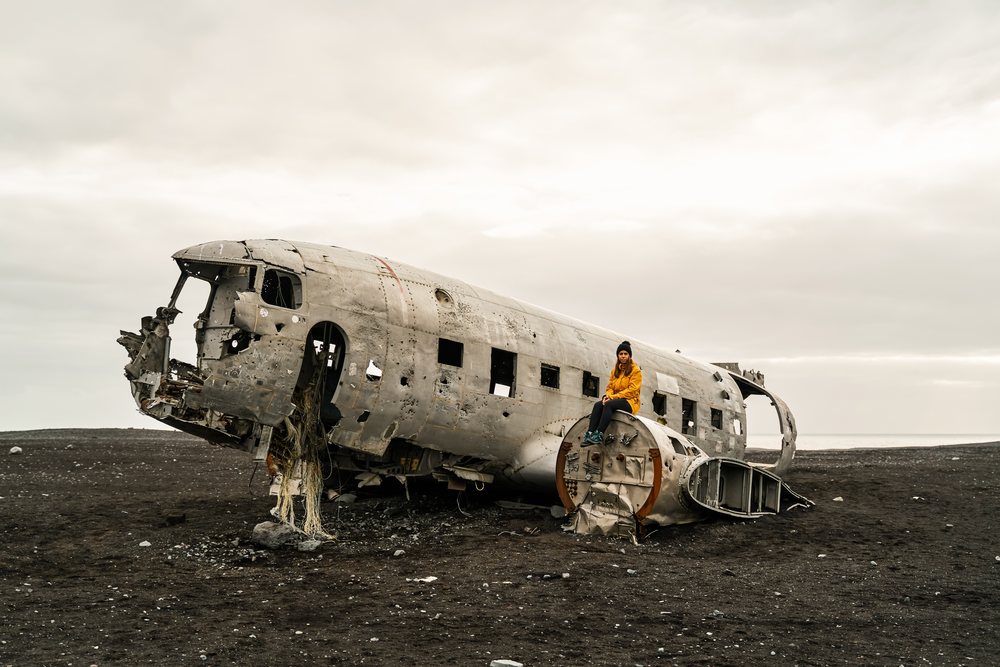 A white woman sits on wreckage of a crashed airplane on a black sand