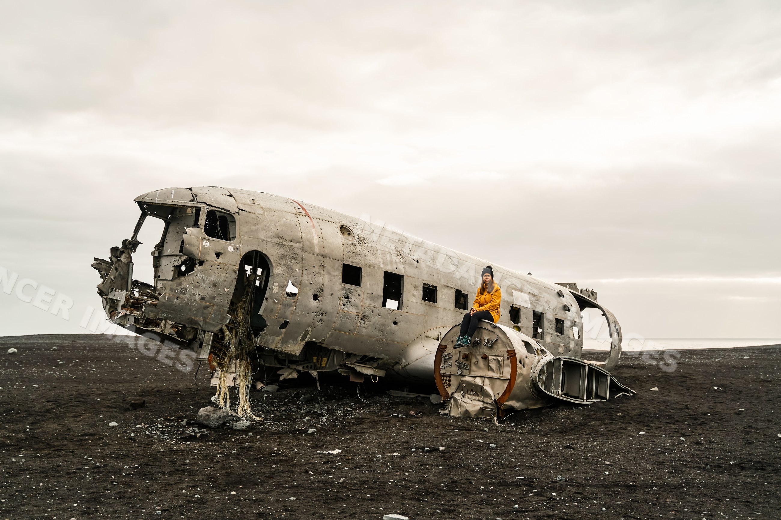 A white woman sits on wreckage of a crashed airplane on a black sand