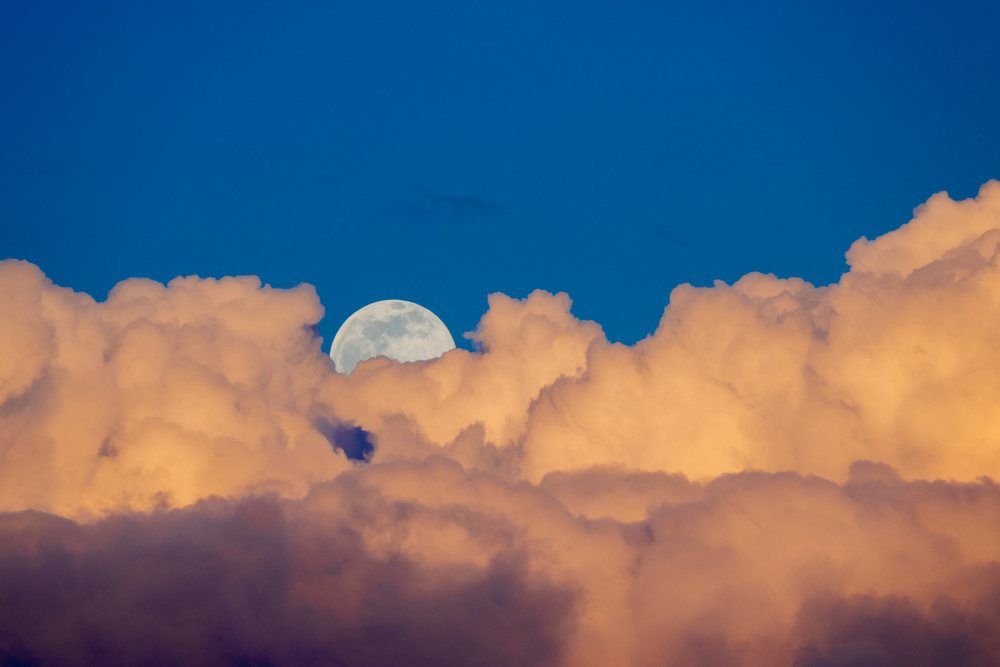 Full moon rising above vibrant orange cumulus clouds in clear blue sky