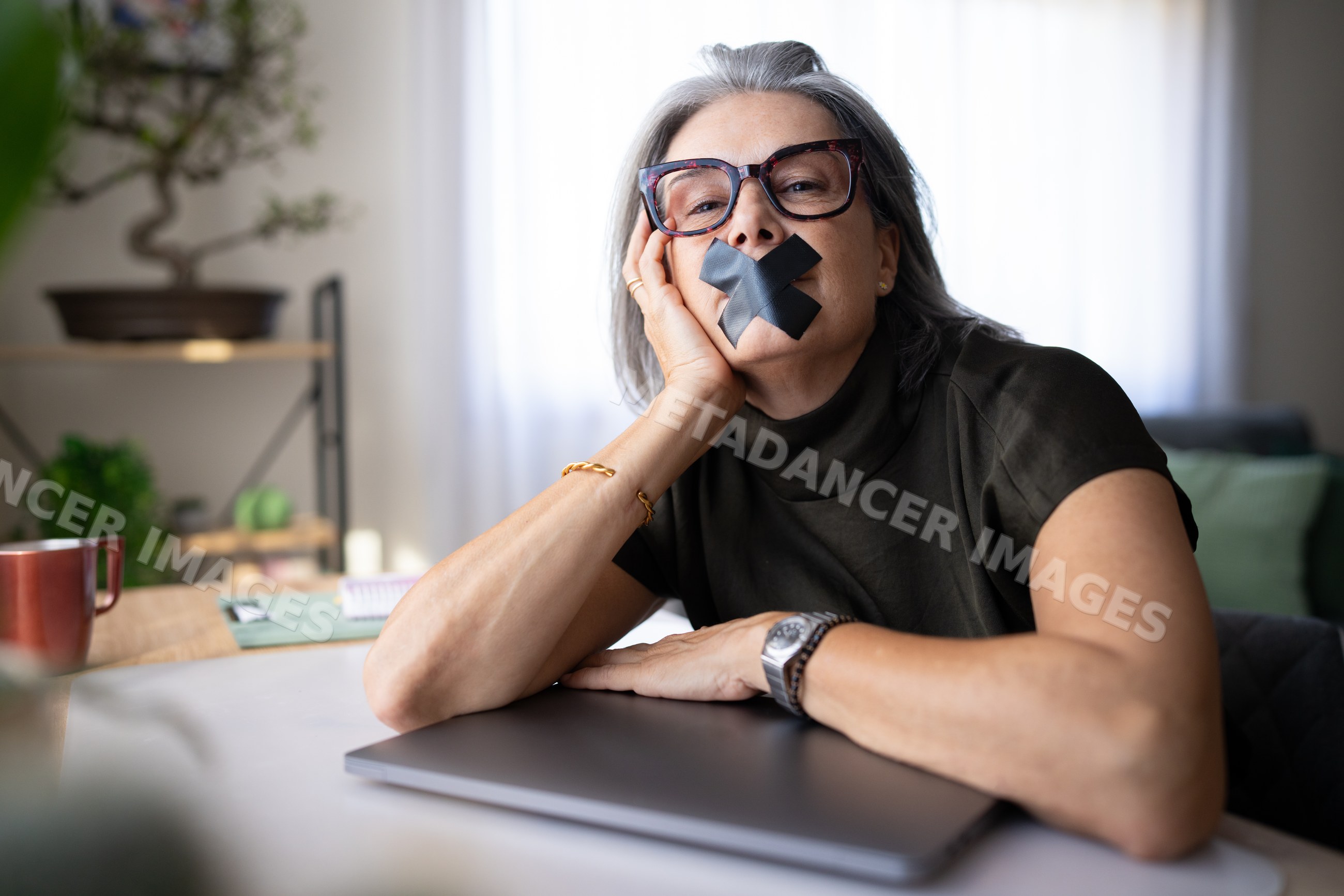 White woman with duct tape over mouth sitting at table indoors