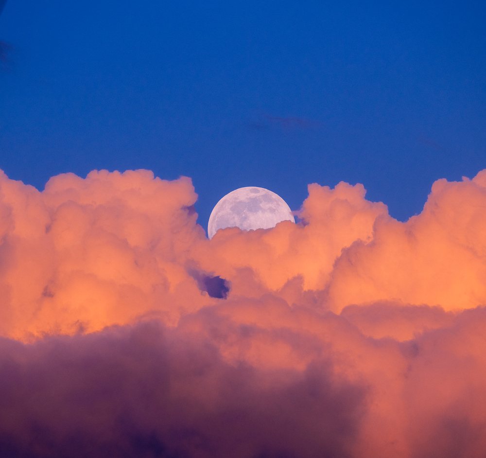 Full moon rising behind vibrant orange clouds at sunset
