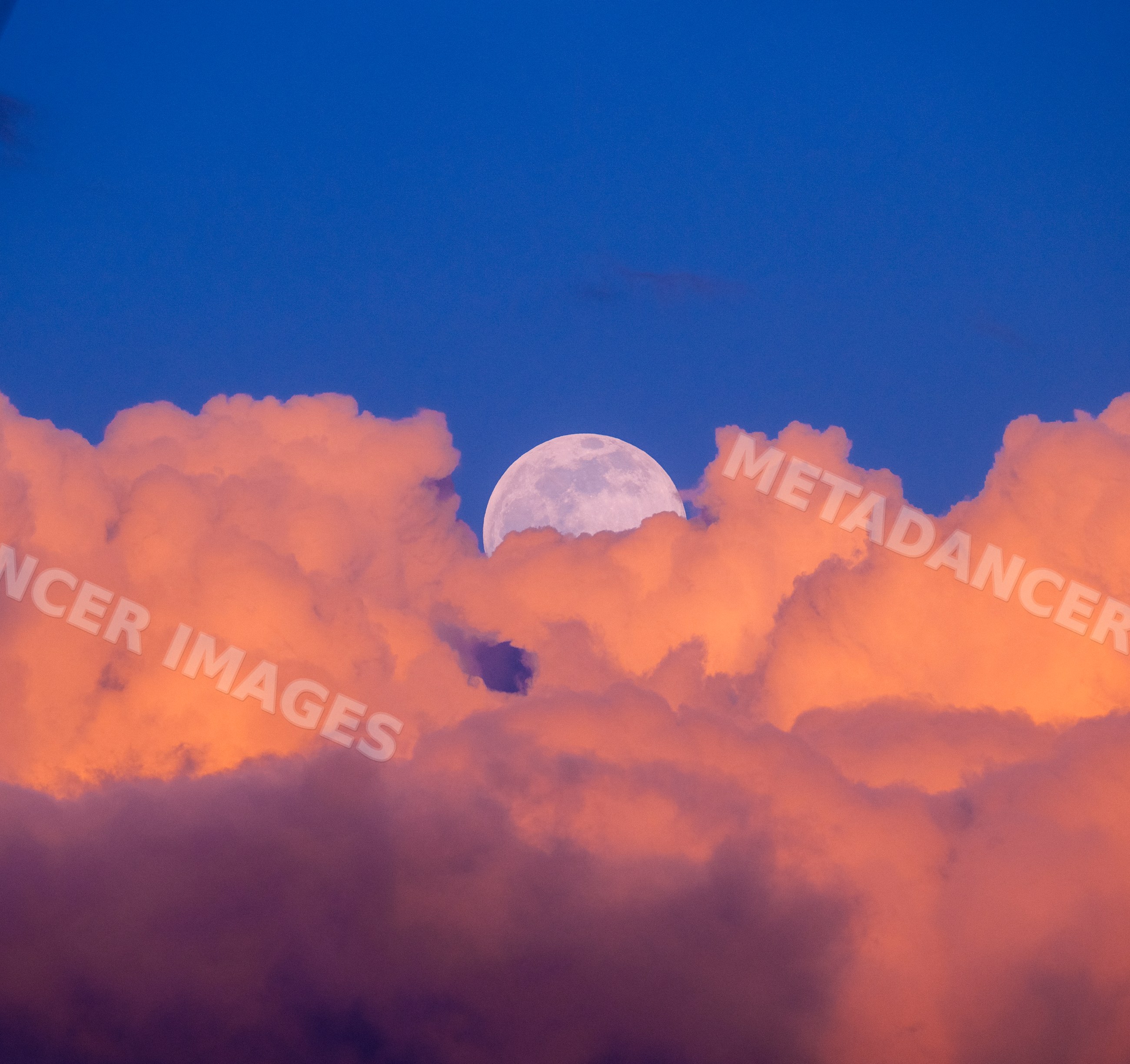 Full moon rising behind vibrant orange clouds at sunset