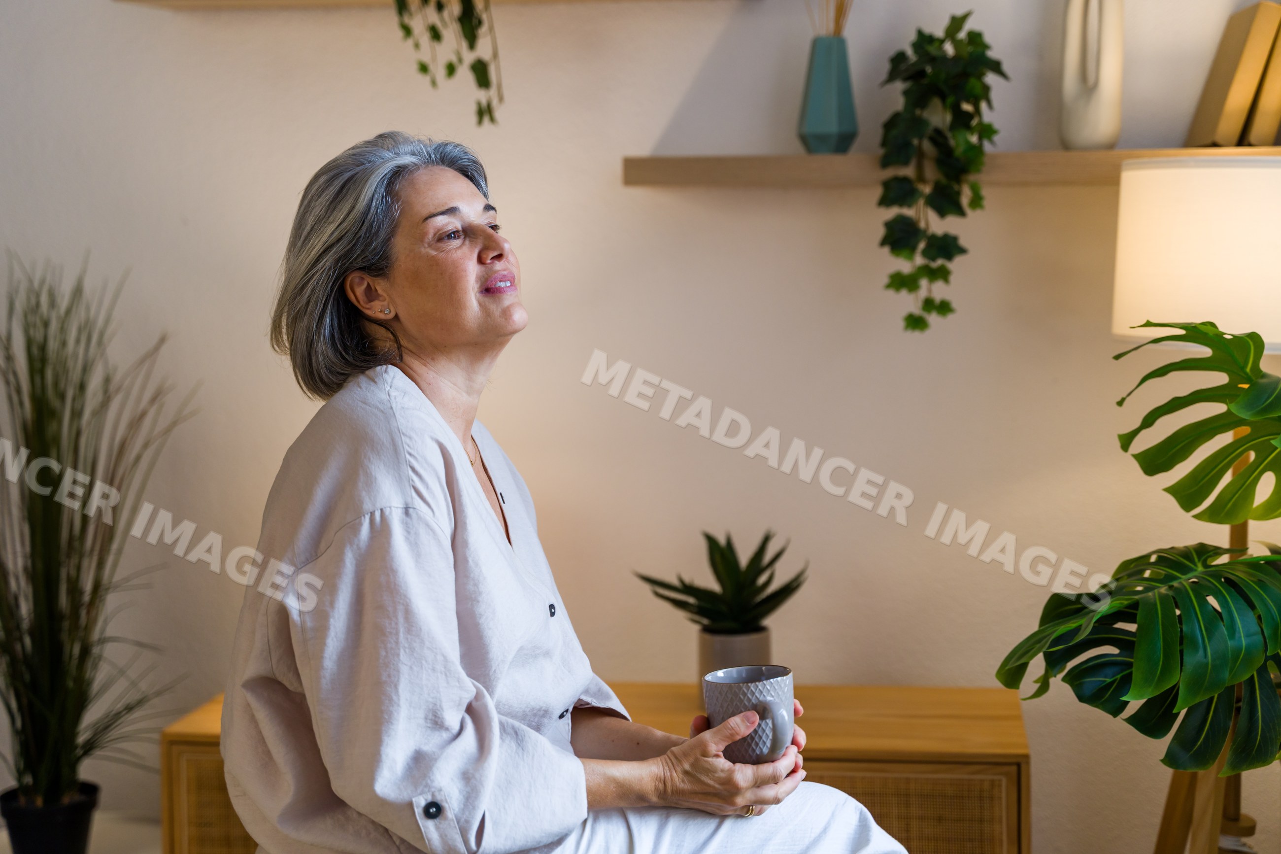 White woman sitting indoors holding mug and thinking