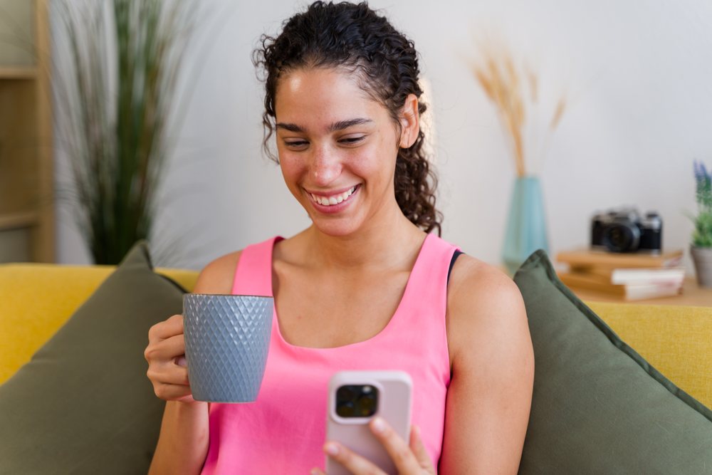 A Hispanic woman smiles while holding a mug and using her smartphone