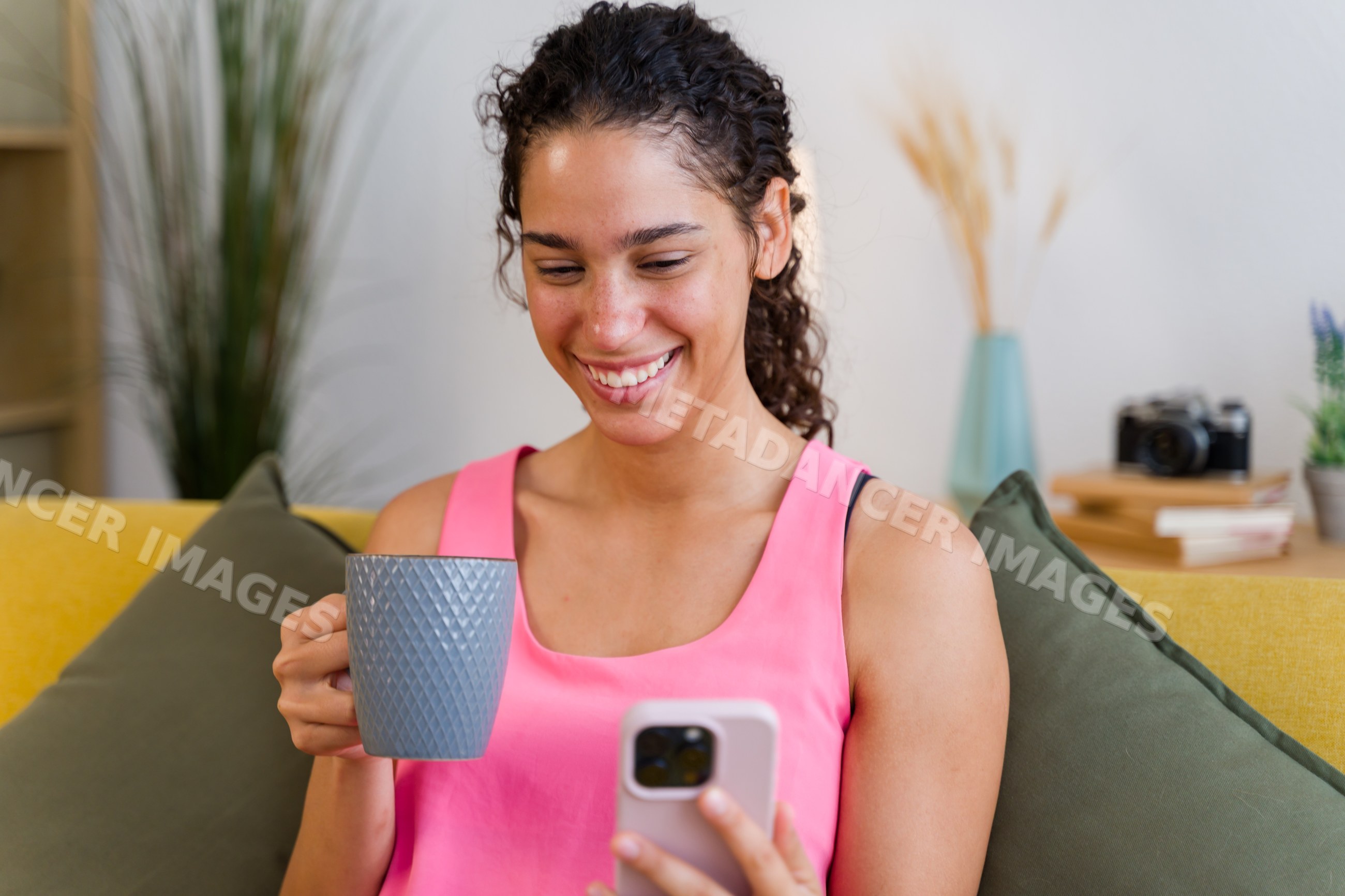 A Hispanic woman smiles while holding a mug and using her smartphone