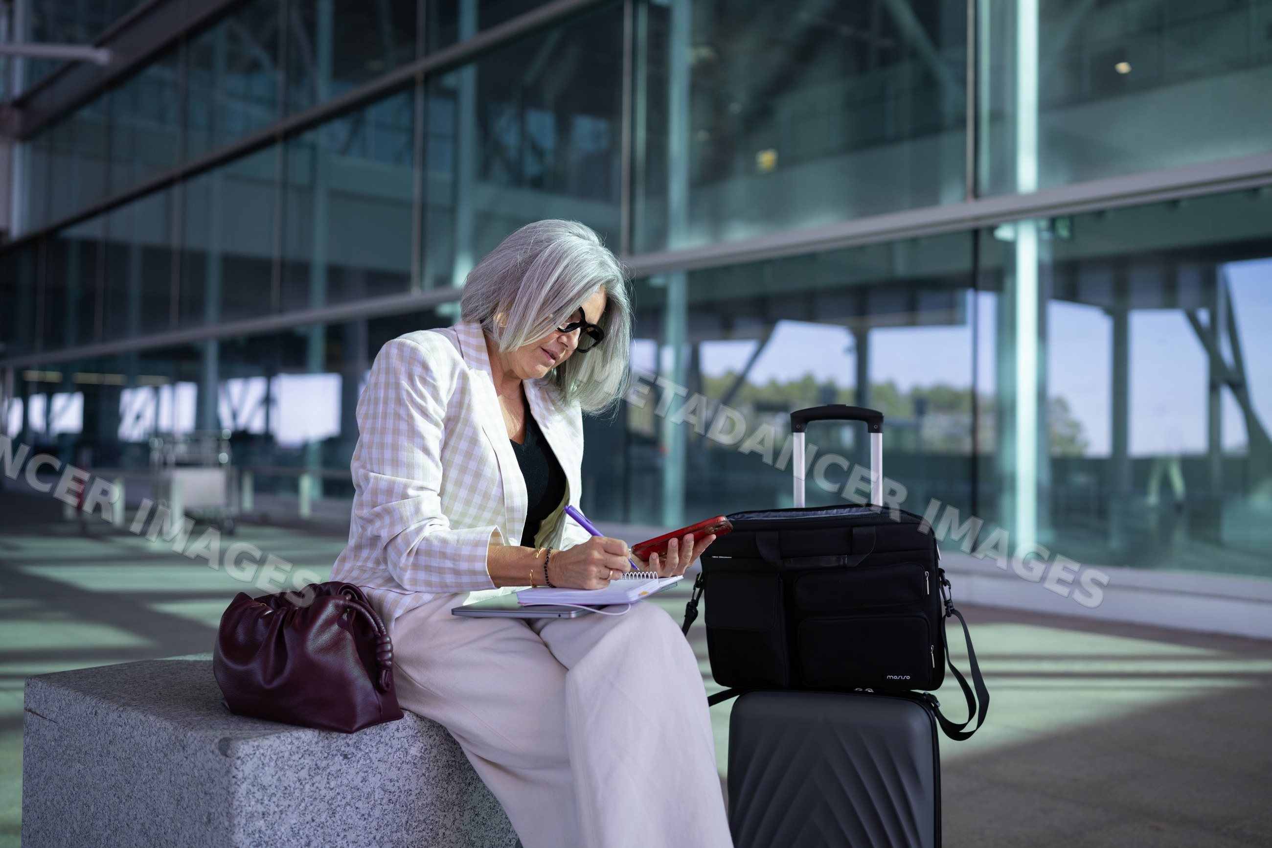 Businesswoman writing in notebook with smartphone by luggage outdoors