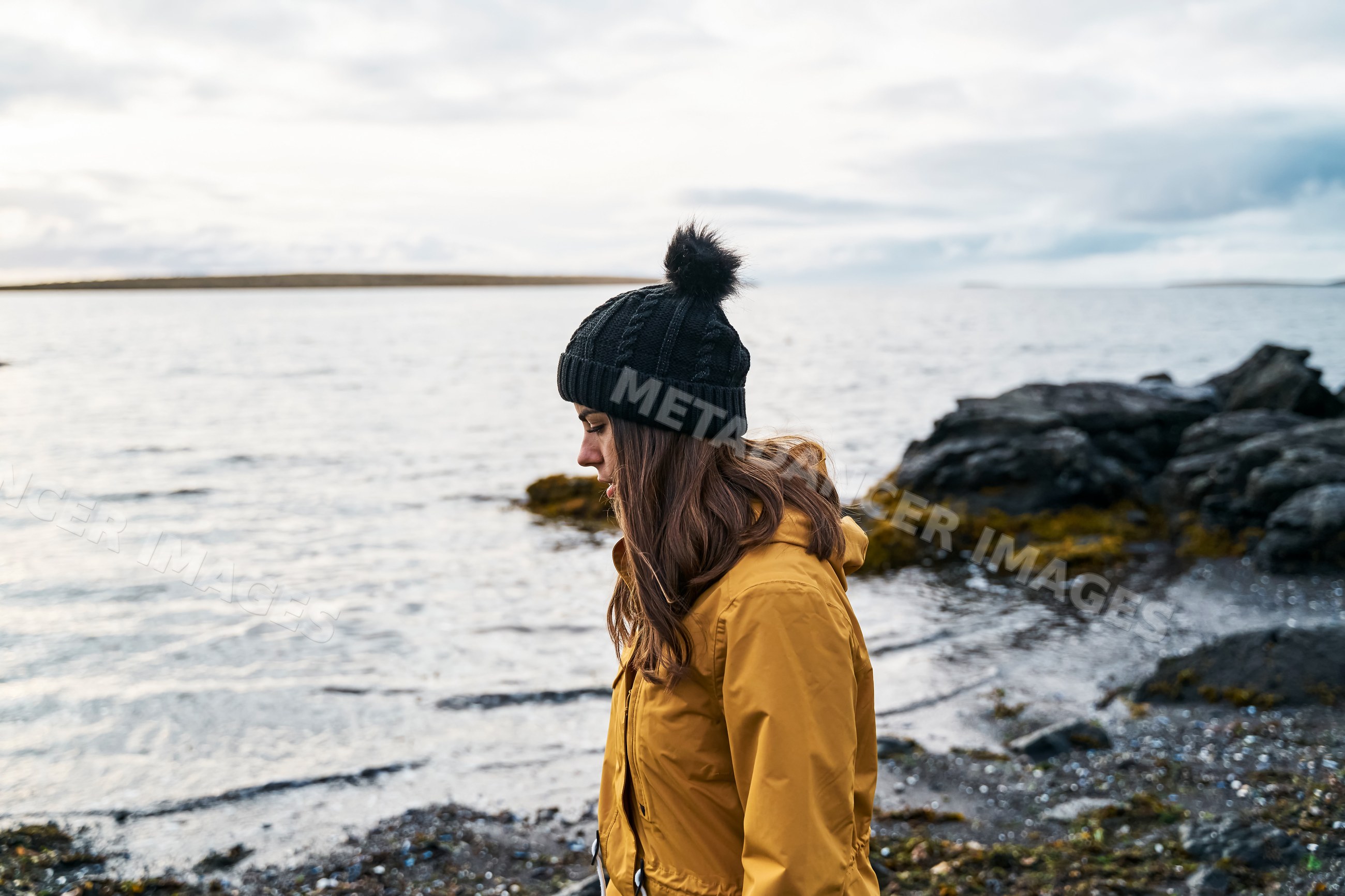 A white woman in a yellow jacket walks along a rocky shore