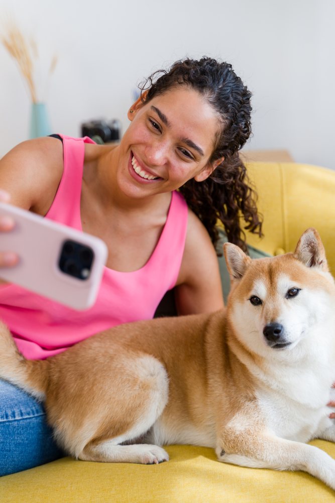 A Hispanic woman happily takes a selfie with her dog on a yellow couch