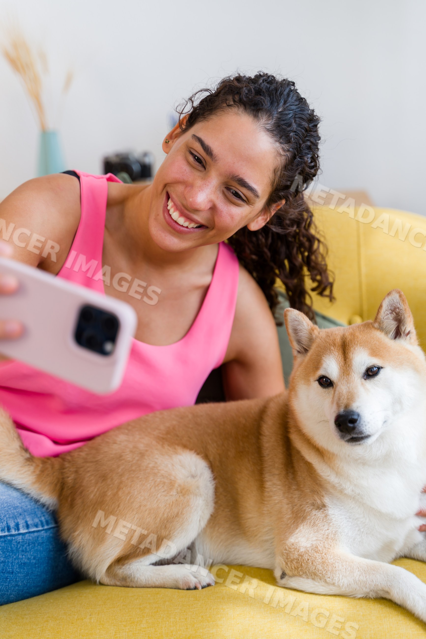 A Hispanic woman happily takes a selfie with her dog on a yellow couch