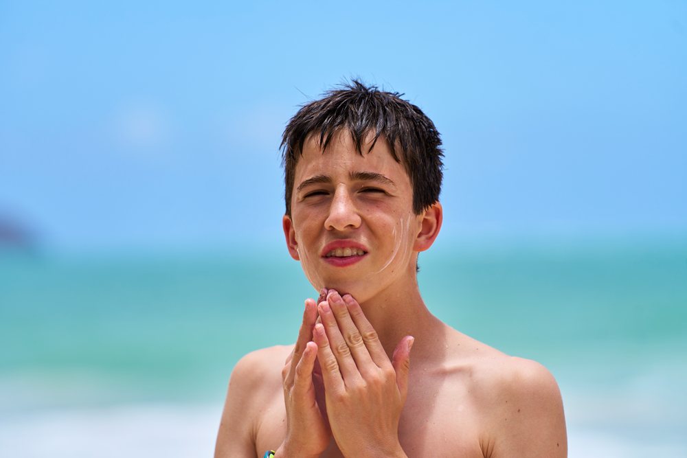 Teenage white boy applying sunscreen on face by ocean in a beach