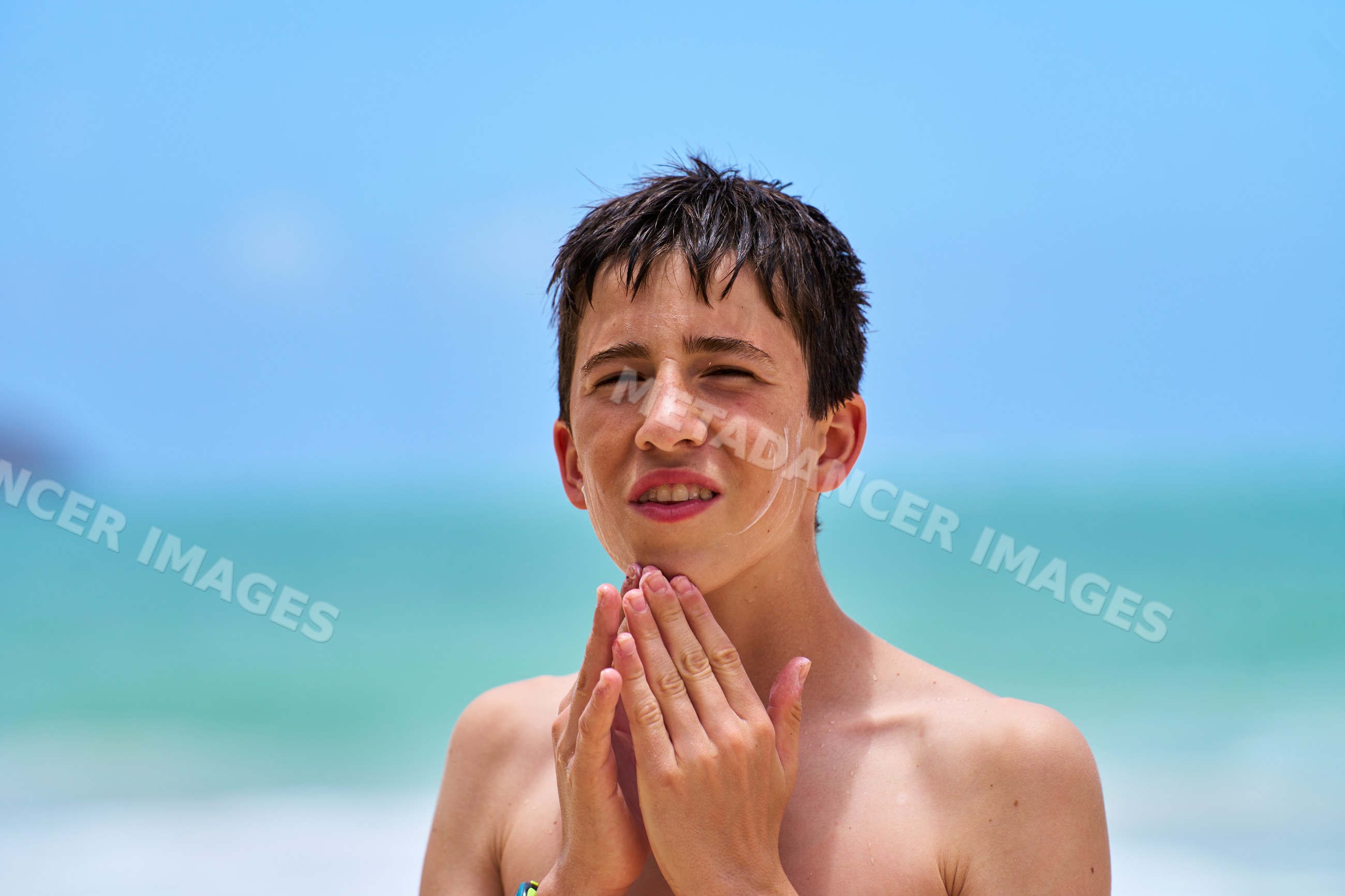 Teenage white boy applying sunscreen on face by ocean in a beach
