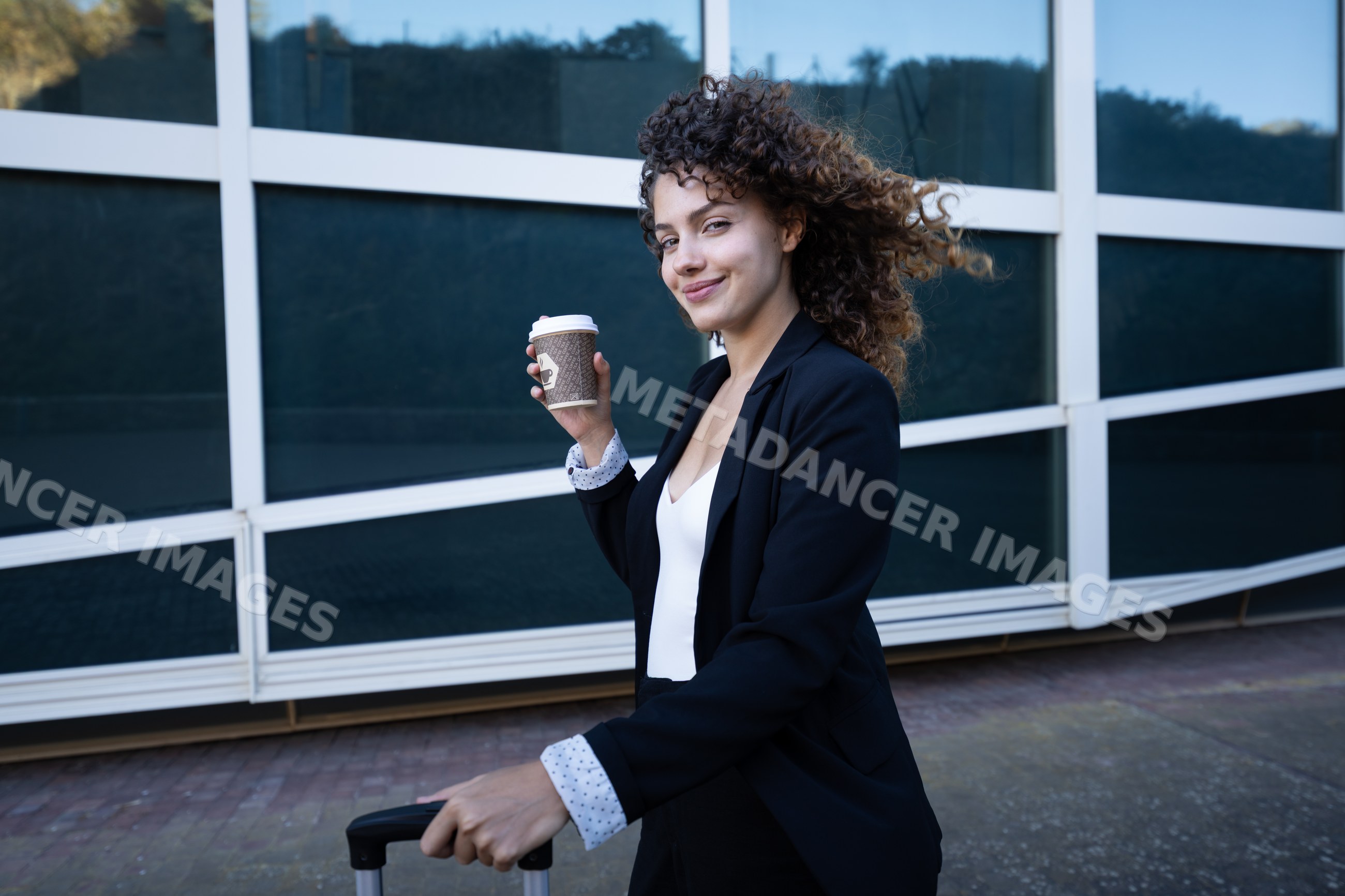 Businesswoman holding coffee cup with suitcase outside modern building