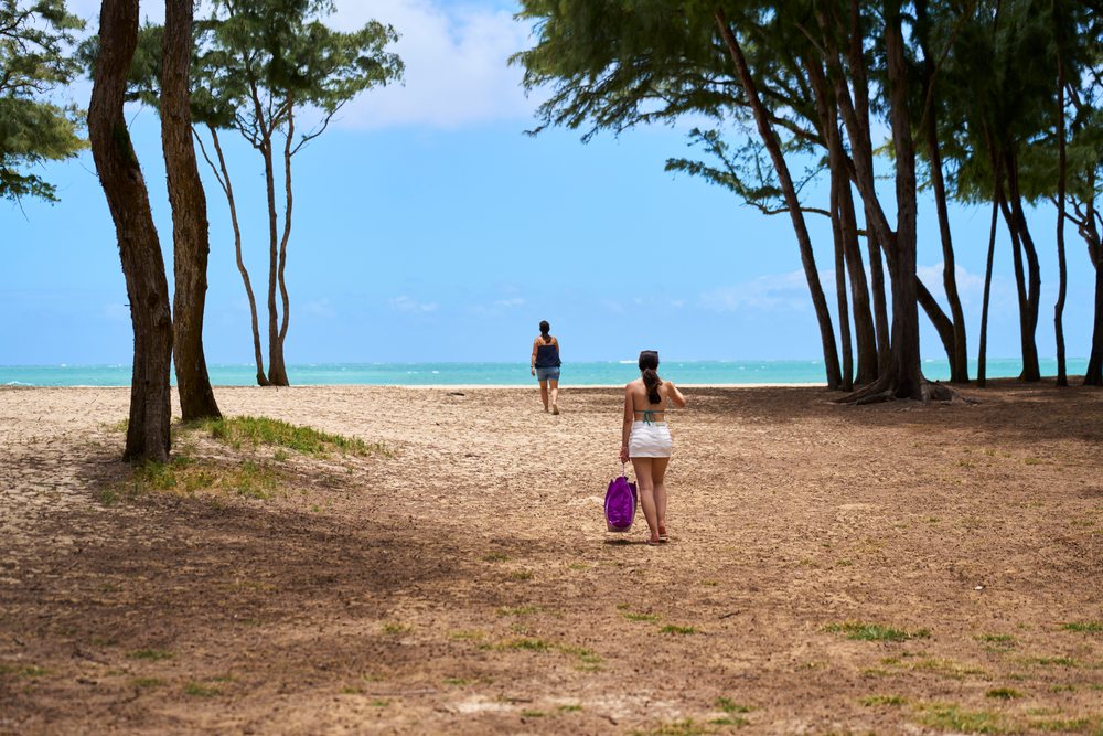 White women walking on a sandy beach path through trees toward ocean