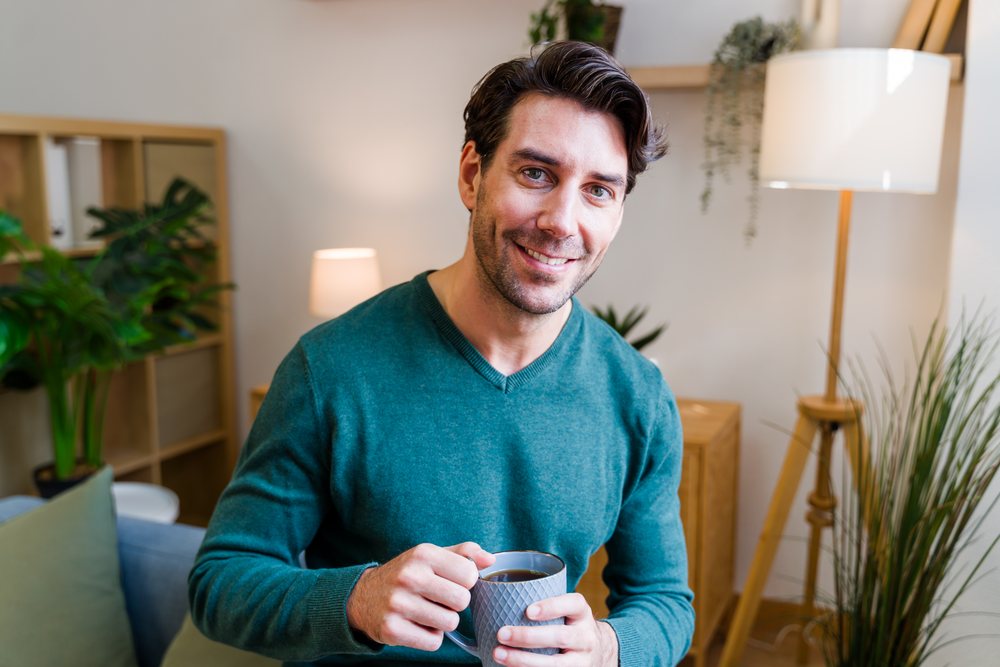 White man holding gray ceramic mug smiling indoors in cozy living room
